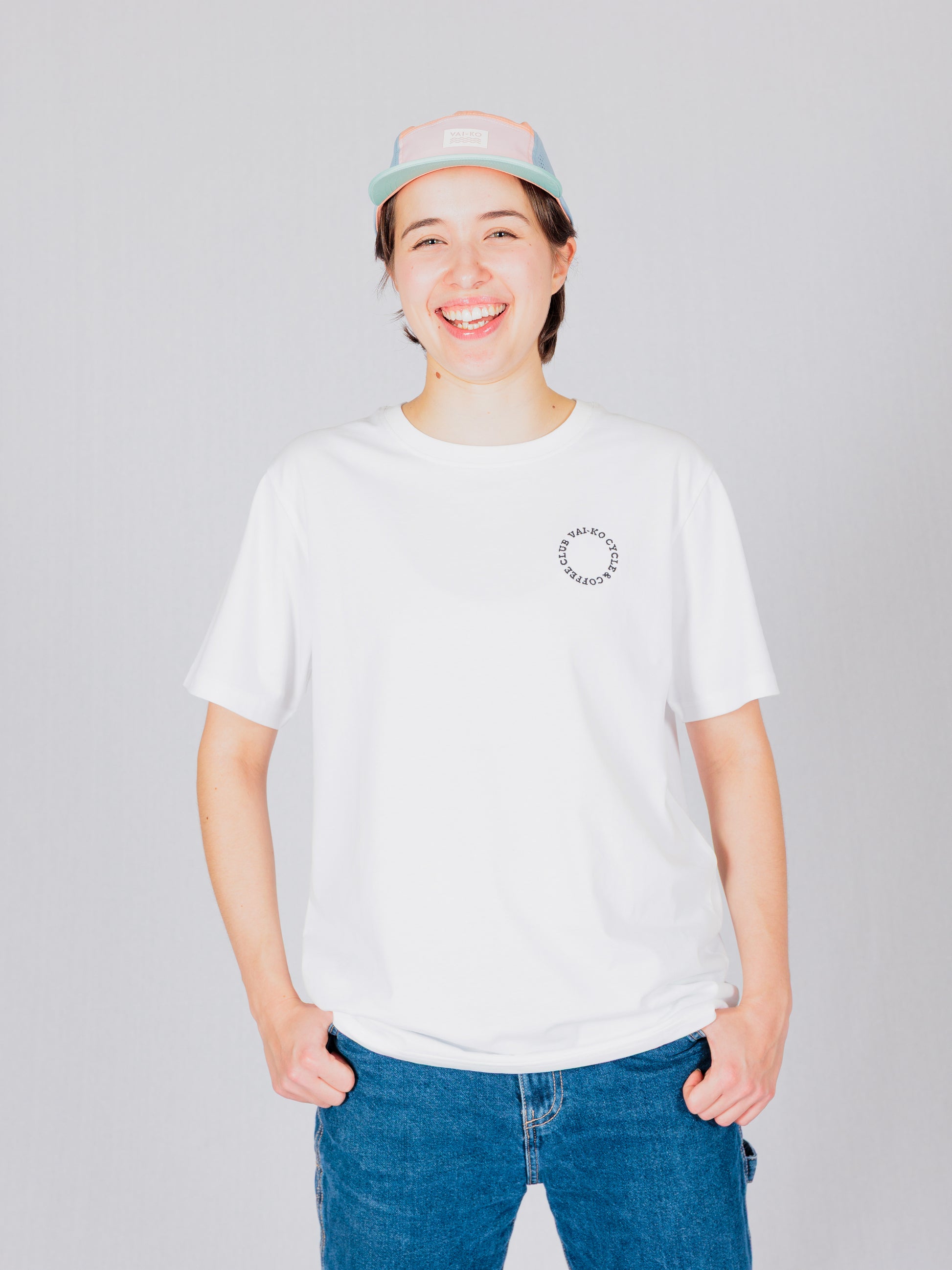 Person in a white t-shirt and pastel cap smiling, standing against a light gray background.