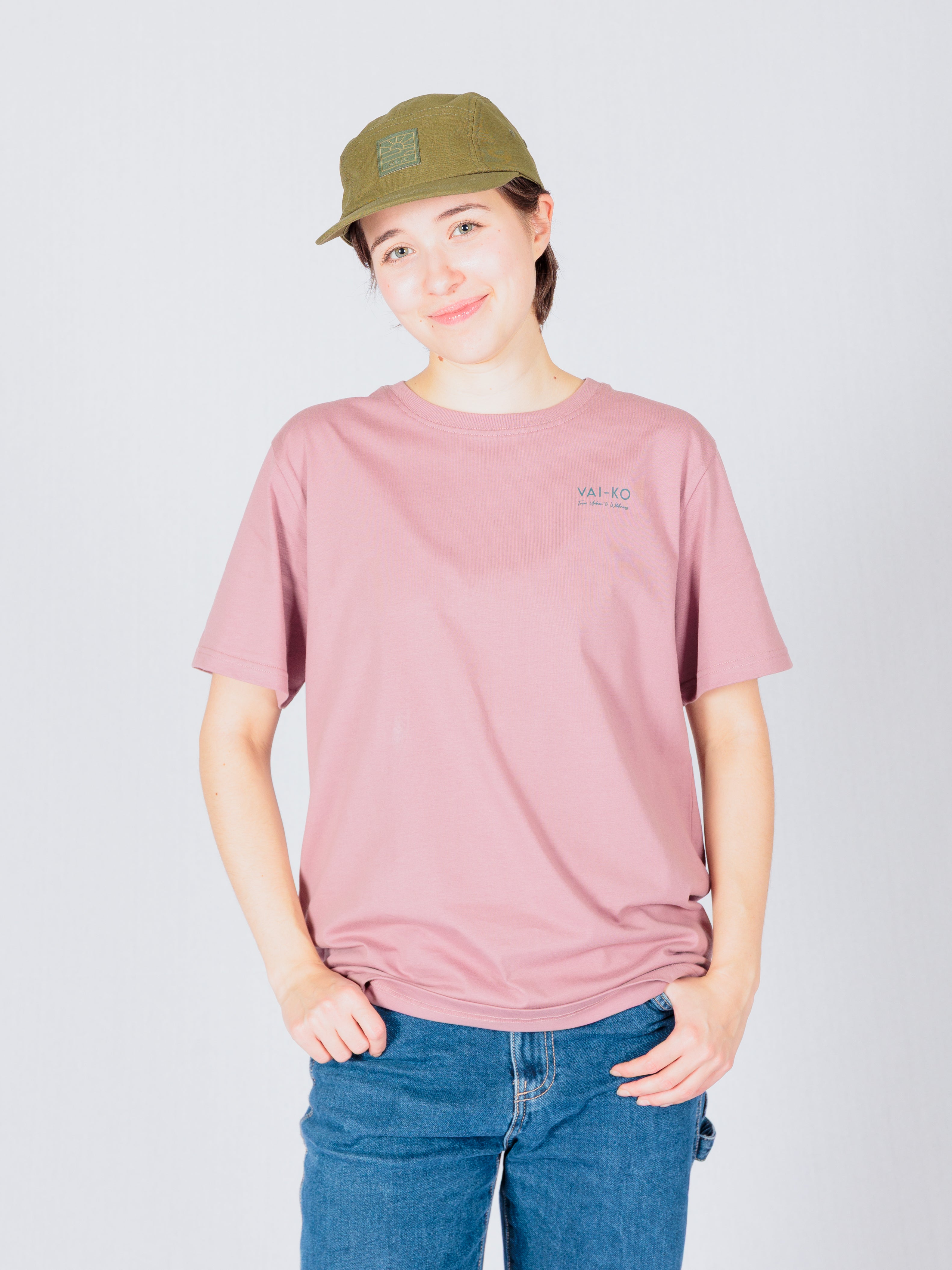 Person wearing a pink t-shirt, green cap, and blue jeans, standing and smiling against a light background.