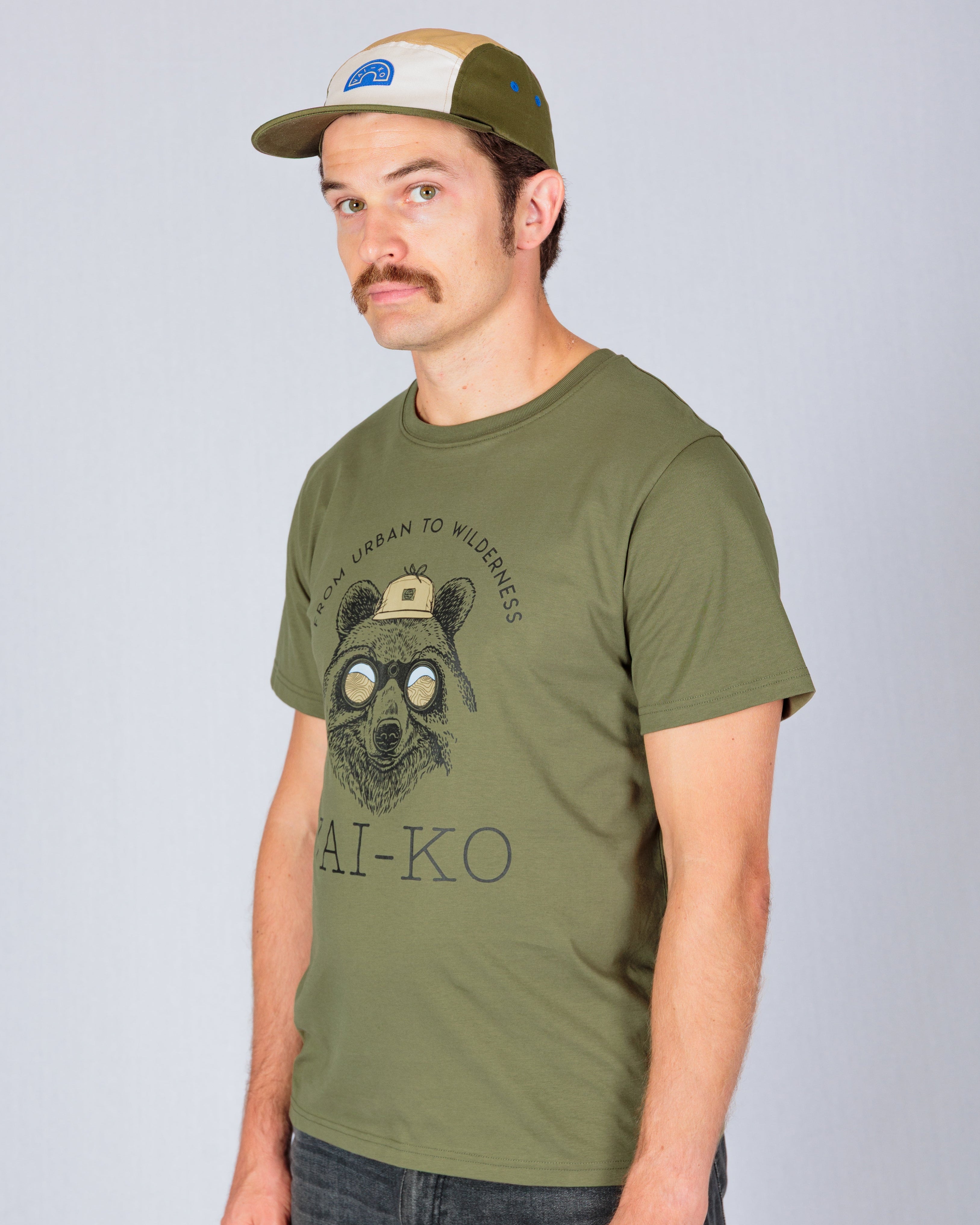 Man in an olive green t-shirt with a bear graphic and a green and beige cap stands against a plain background.