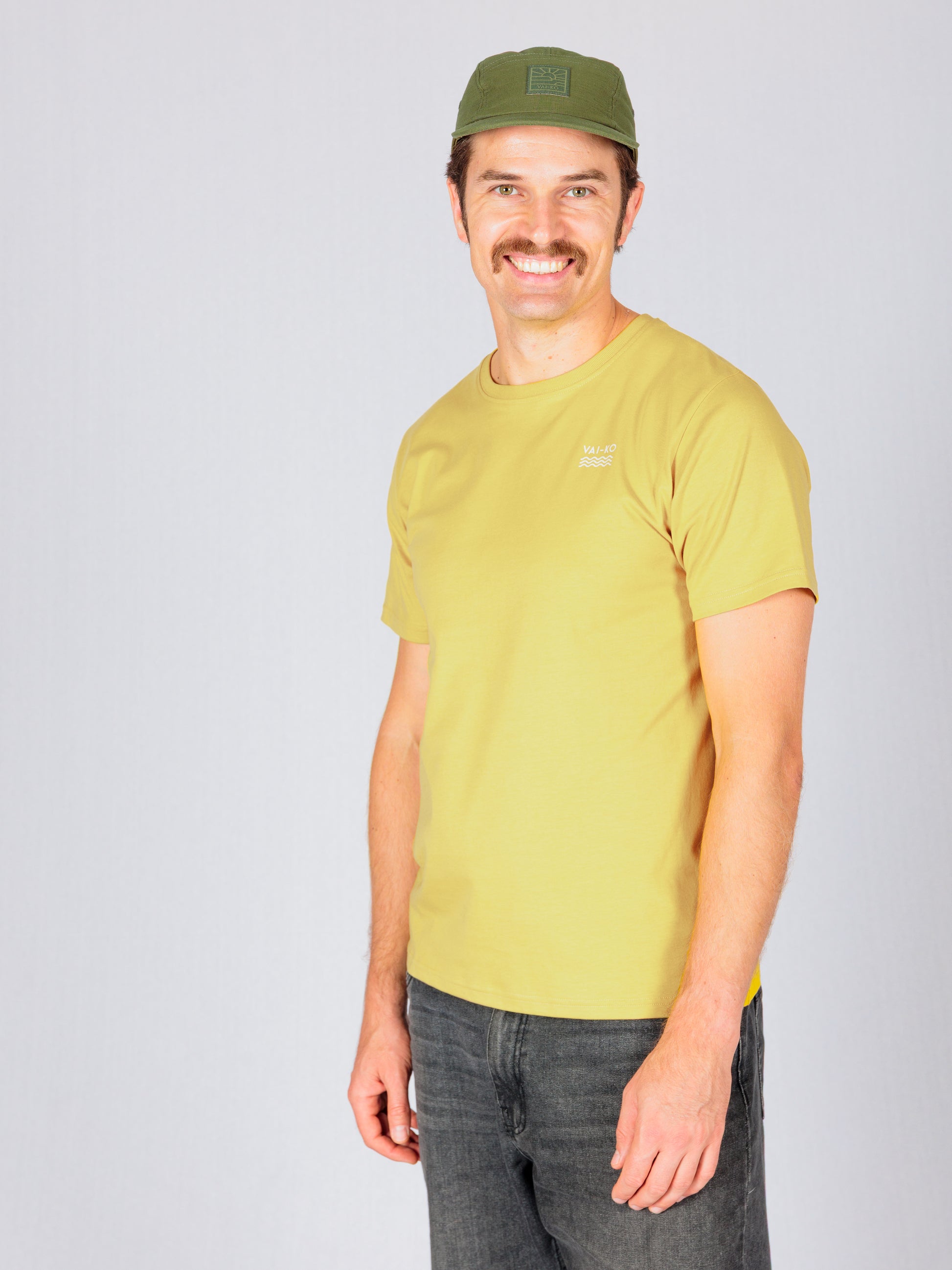 Man in a yellow t-shirt, green cap, and black jeans smiling against a plain light background.