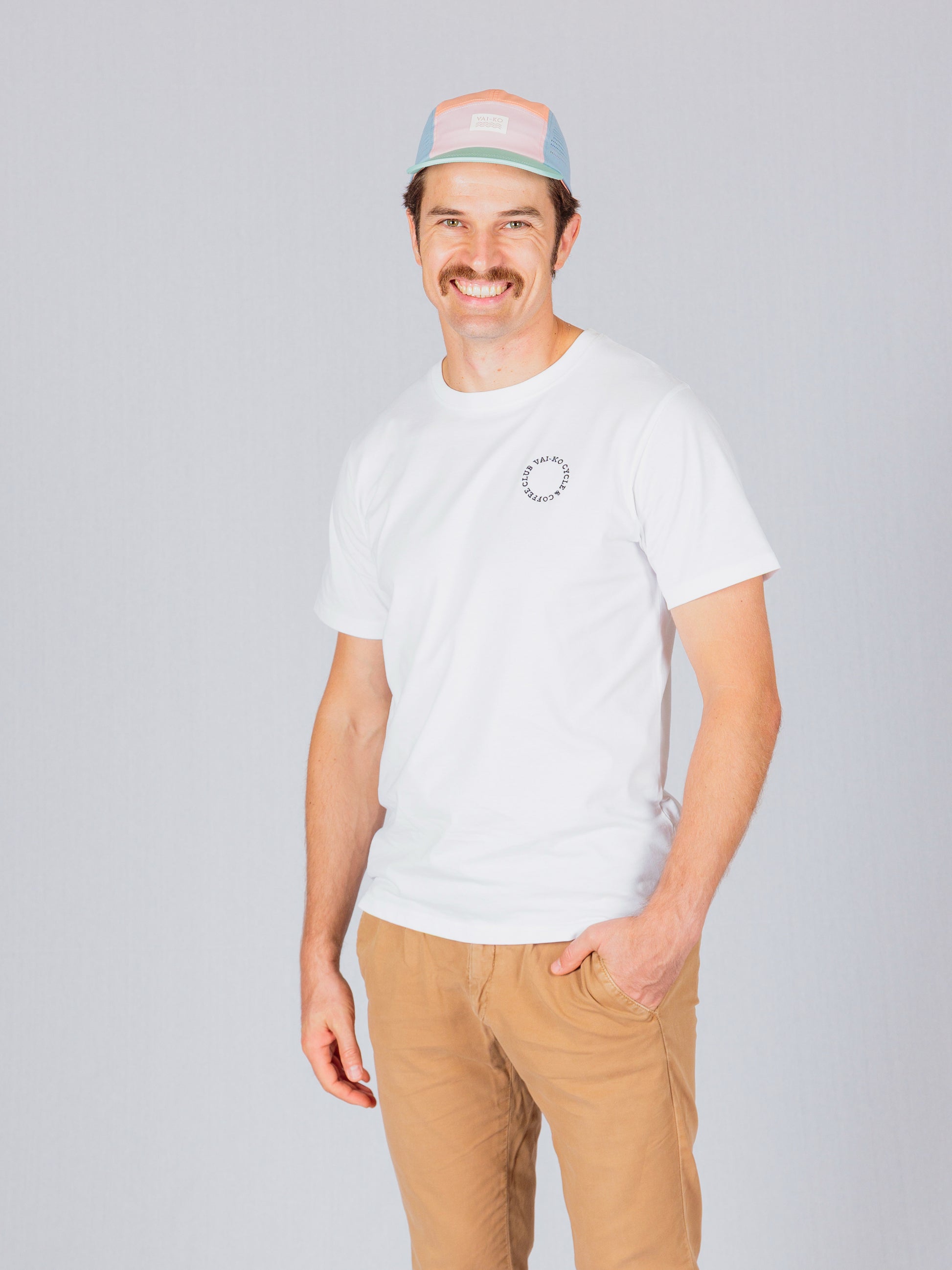 Smiling man in a white t-shirt, khaki pants, and a pastel cap standing against a light gray background.