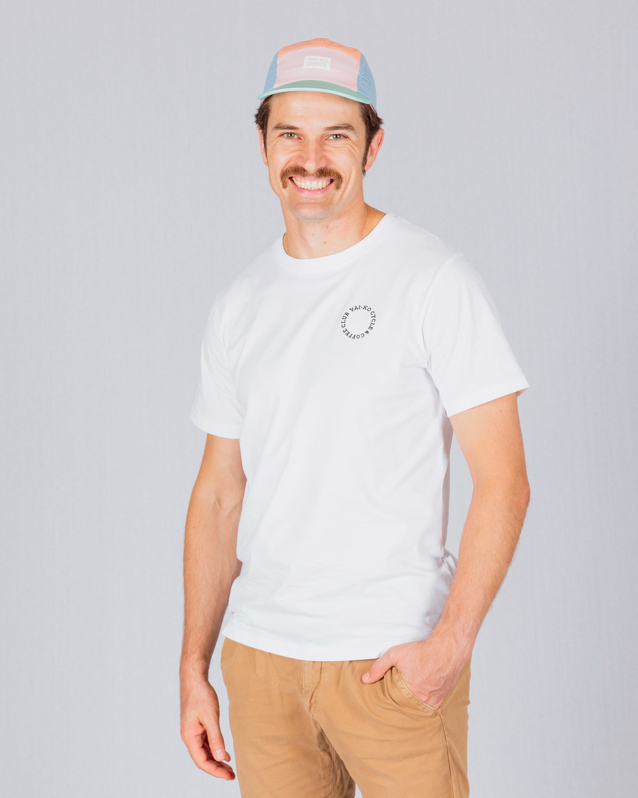 Smiling man in a white t-shirt, khaki pants, and a pastel cap standing against a light gray background.