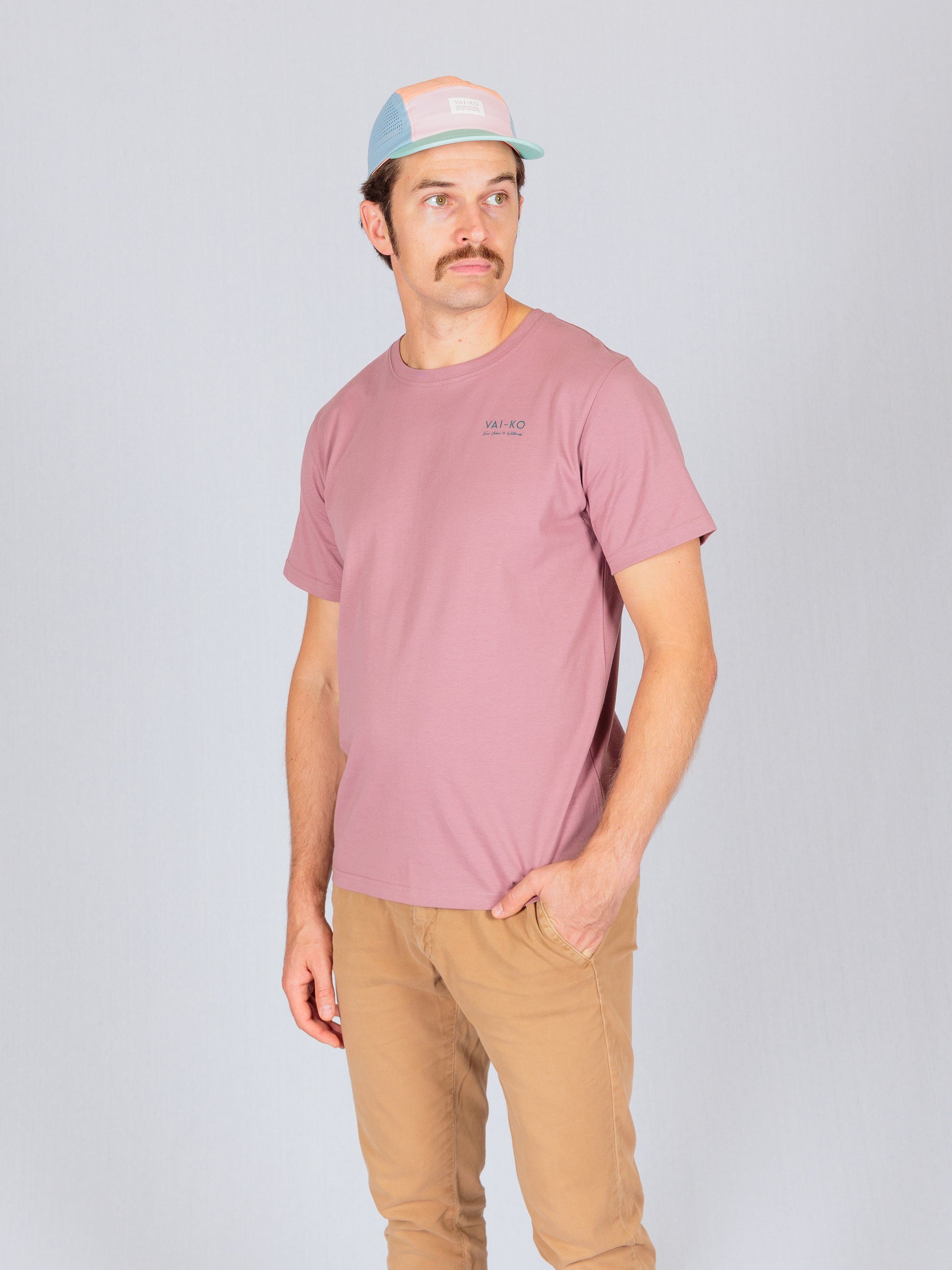 Man in a pink t-shirt, tan pants, and pastel cap stands against a plain light gray background.