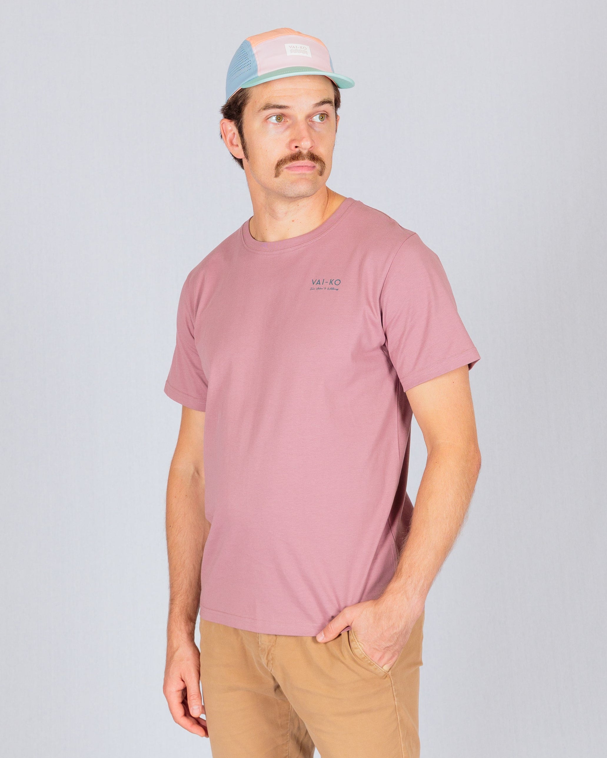 Man in a pink t-shirt, tan pants, and pastel cap stands against a plain light gray background.