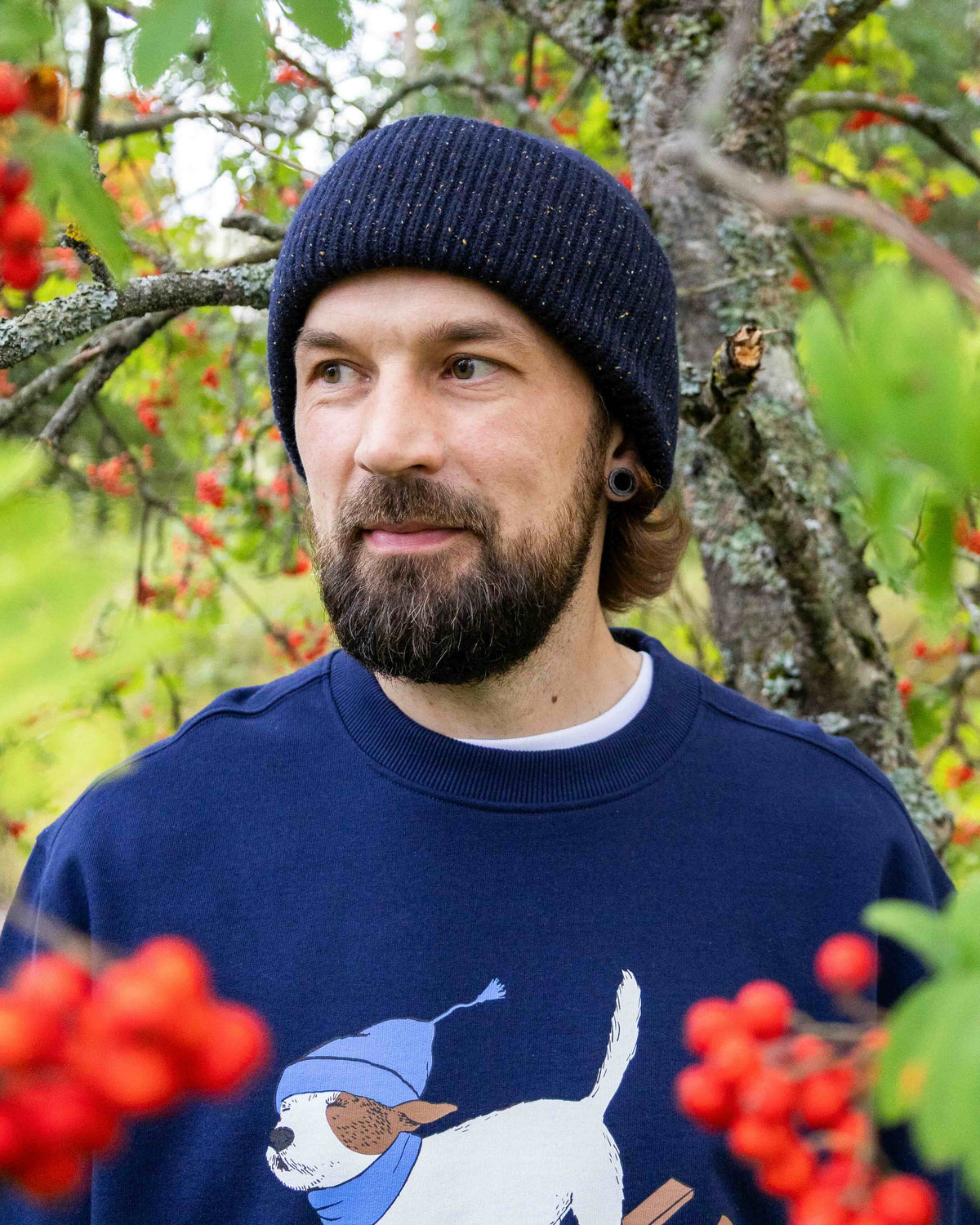 Man with a beard in a navy beanie and dog sweatshirt stands among branches with red berries.