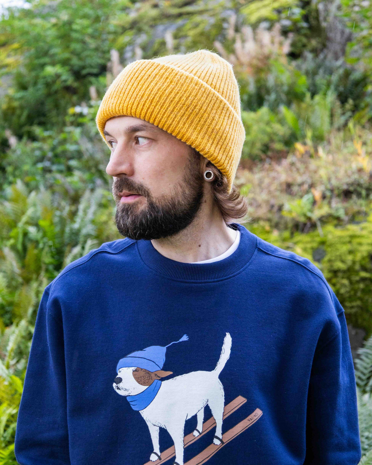 Bearded person in yellow beanie and blue sweater with dog graphic, standing outdoors by greenery.