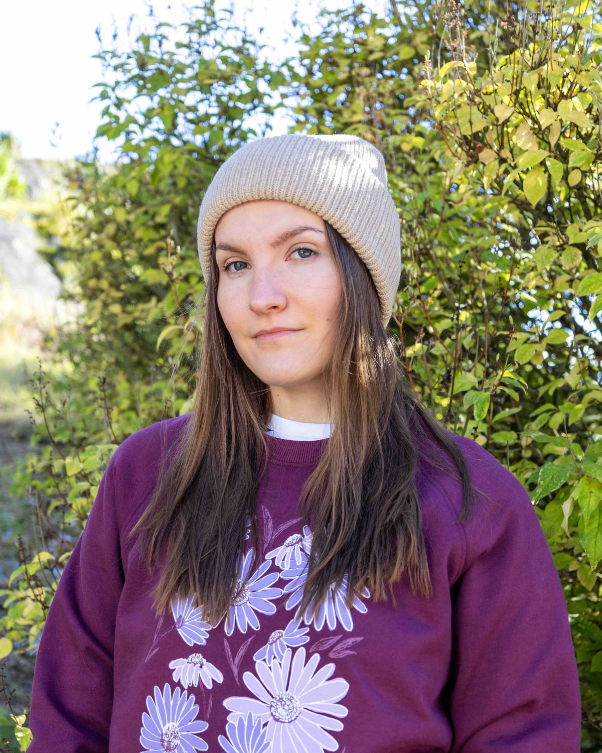 Woman in beige knit hat and purple sweatshirt with flower design, standing outside by green bushes.