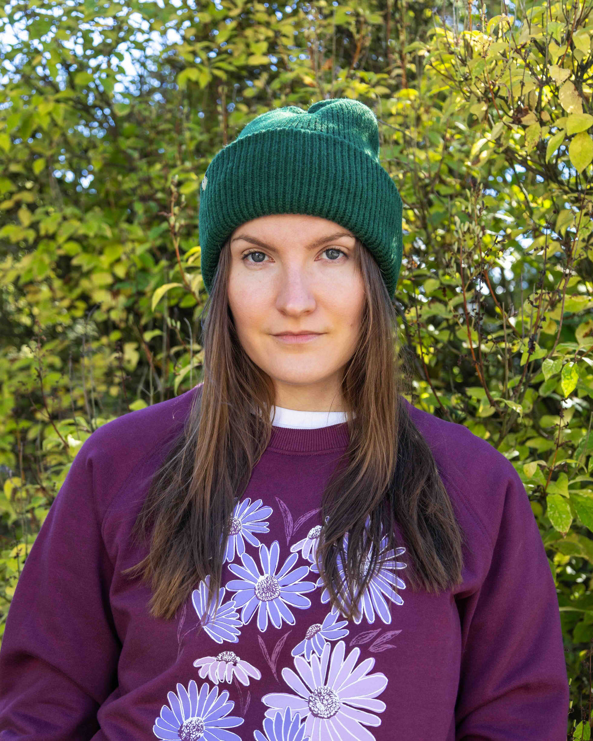 Woman in a green beanie and purple floral sweater stands outside with green foliage in the background.