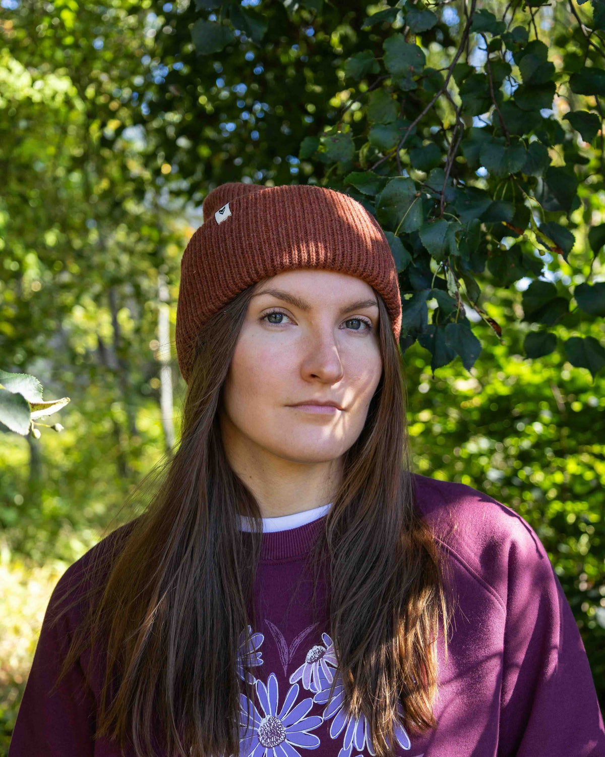 Woman in maroon hat and purple sweatshirt stands outdoors with green foliage in the background.