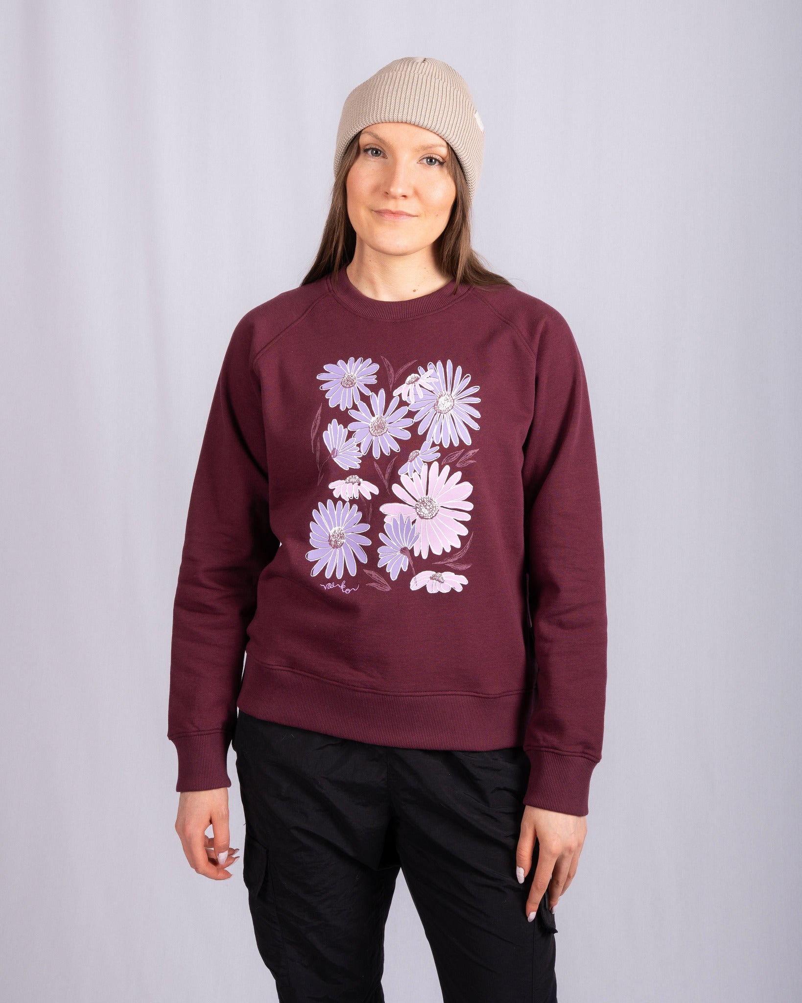 Woman in maroon sweatshirt with purple daisy print, beige beanie, and black pants stands against a gray background.