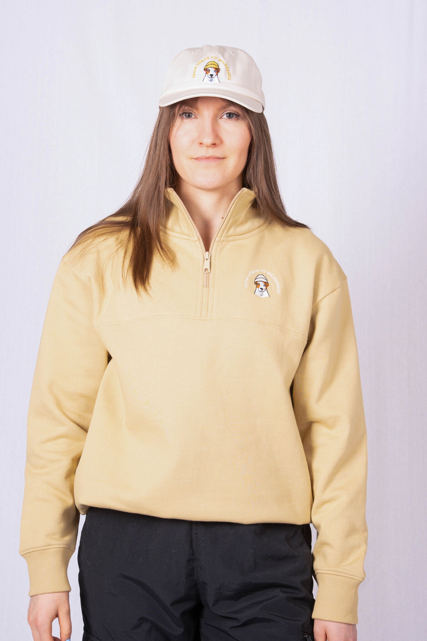 Woman in a beige quarter-zip sweatshirt and matching cap with embroidered logos, standing against a white background.