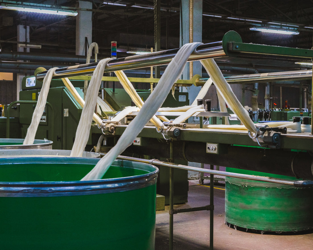 Textile machine feeds white fabric strands into large green containers in a brightly lit industrial factory.