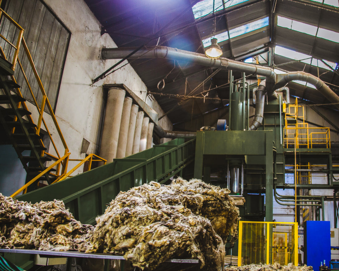 Industrial factory interior with machinery and piles of raw wool under bright overhead lighting.