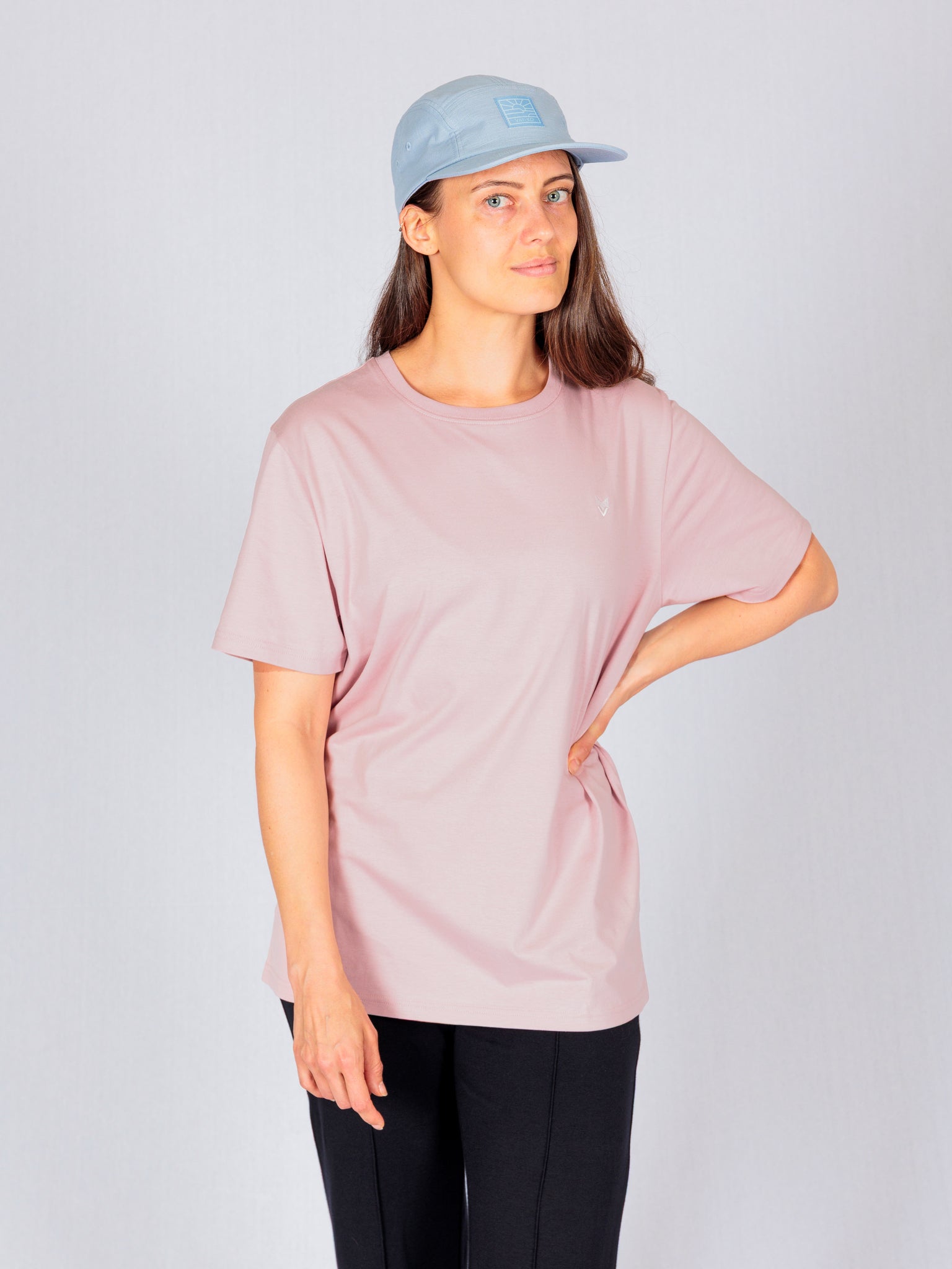 Woman wearing a light blue cap, pink t-shirt, and black pants, standing with one hand on her hip.