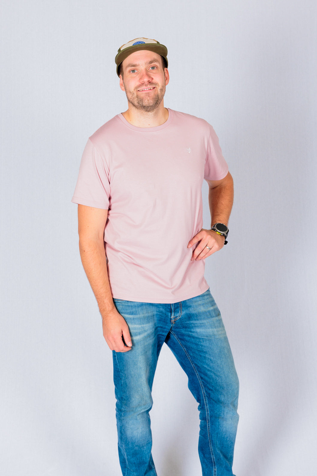 Man in a pink t-shirt, blue jeans, and cap standing and smiling against a plain light background.