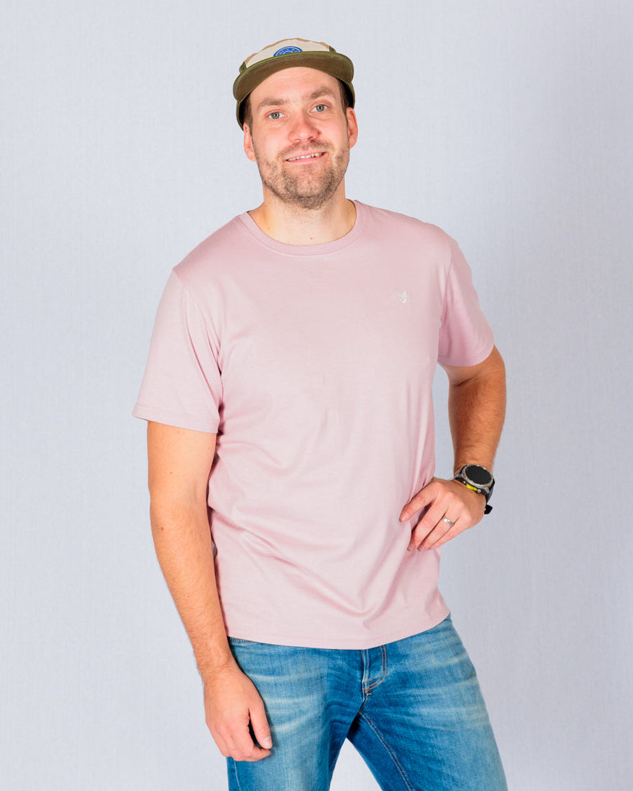 Man in a pink t-shirt, blue jeans, and cap standing and smiling against a plain light background.