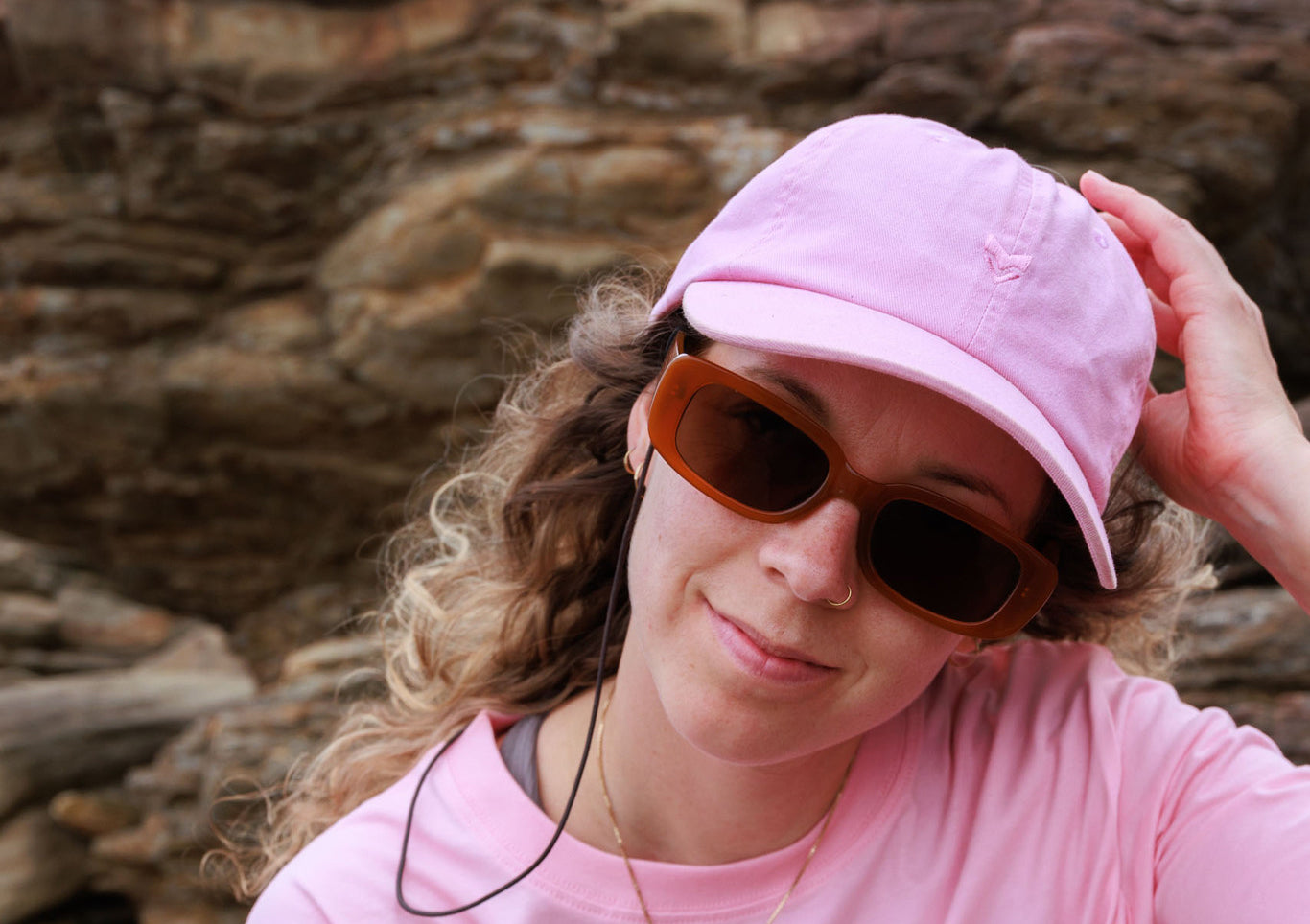 Woman in a pink cap and shirt with sunglasses, smiling and touching her head, rocky background behind her.