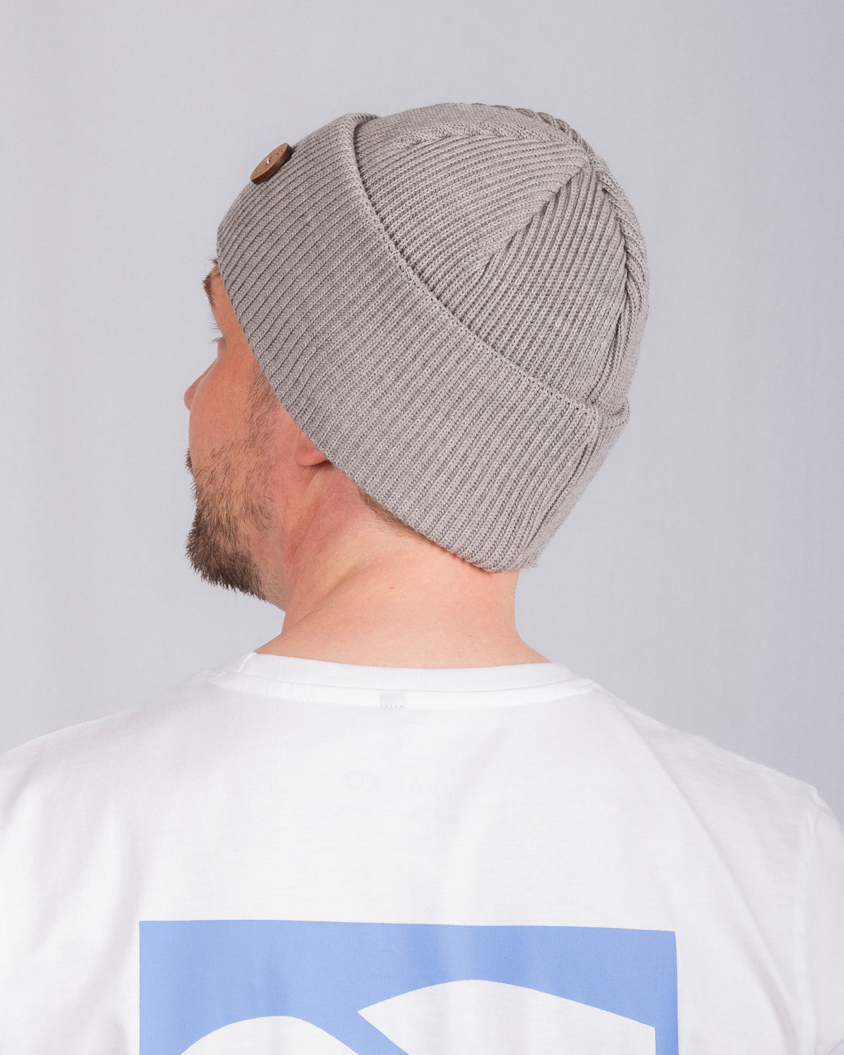 Man in gray knit beanie and white shirt with blue graphic, shown from behind against a plain background.