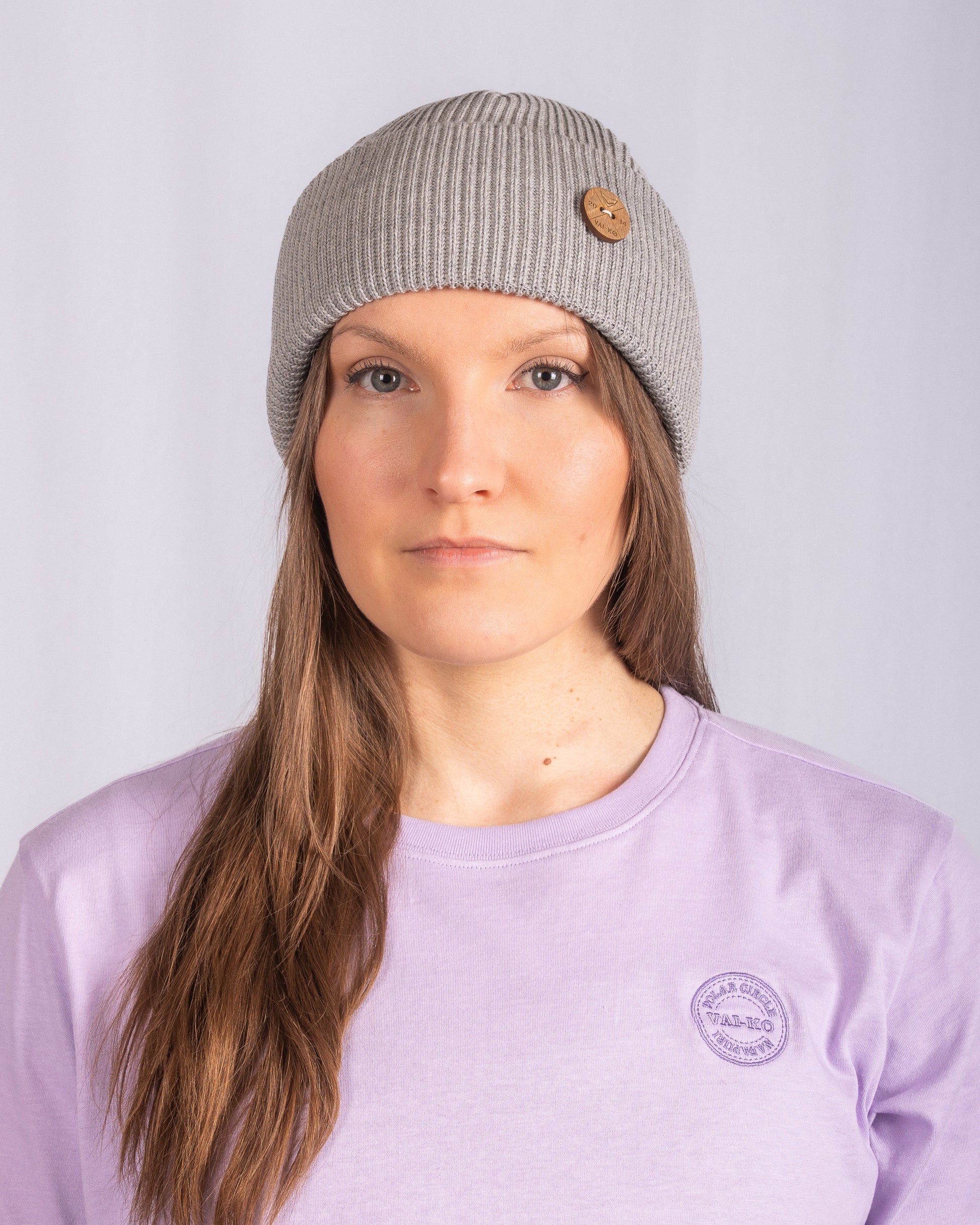A woman wearing a gray knit beanie and a light purple shirt stands against a plain light background.