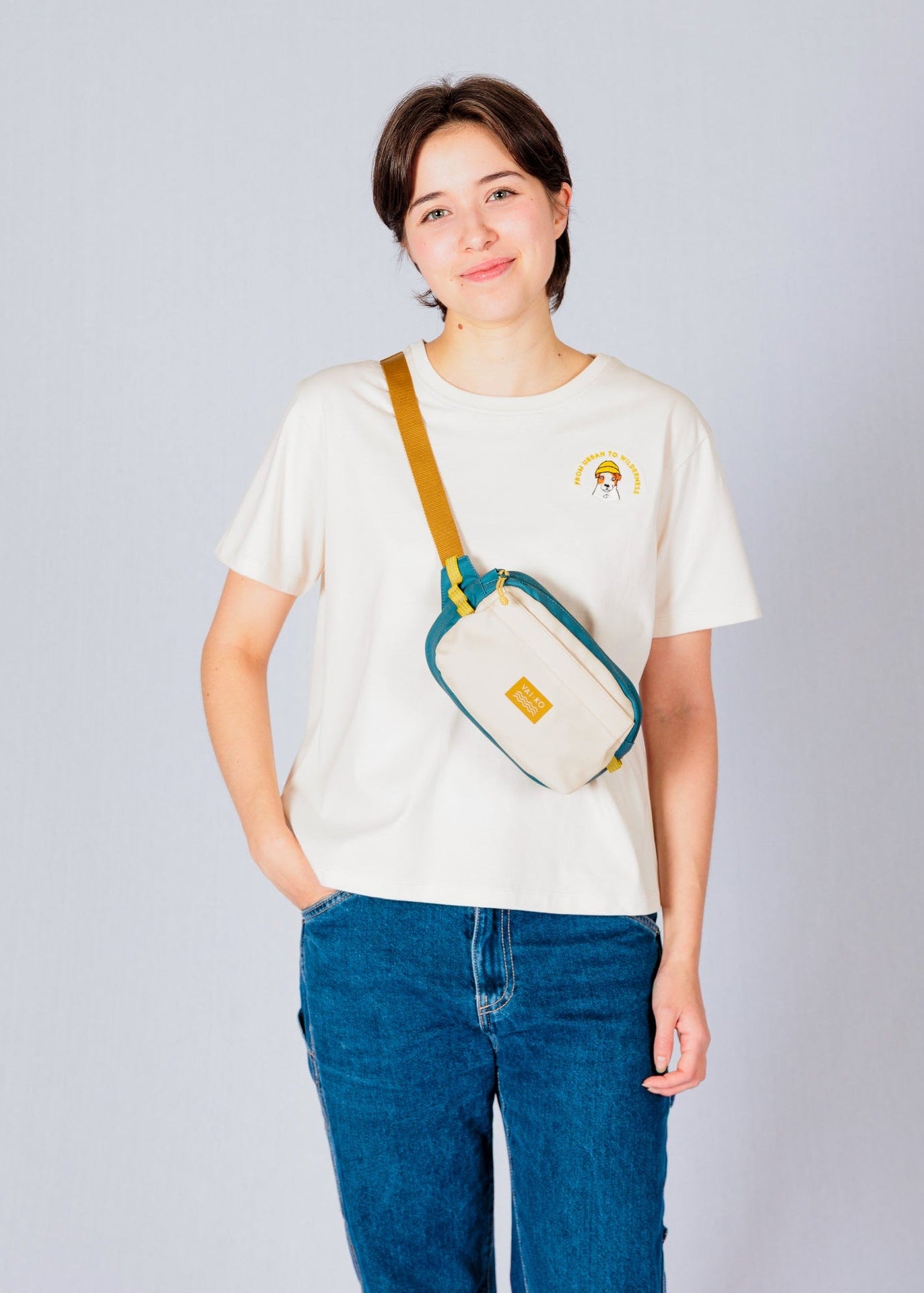 Person in a white t-shirt and jeans, smiling, with a small crossbody bag against a plain background.