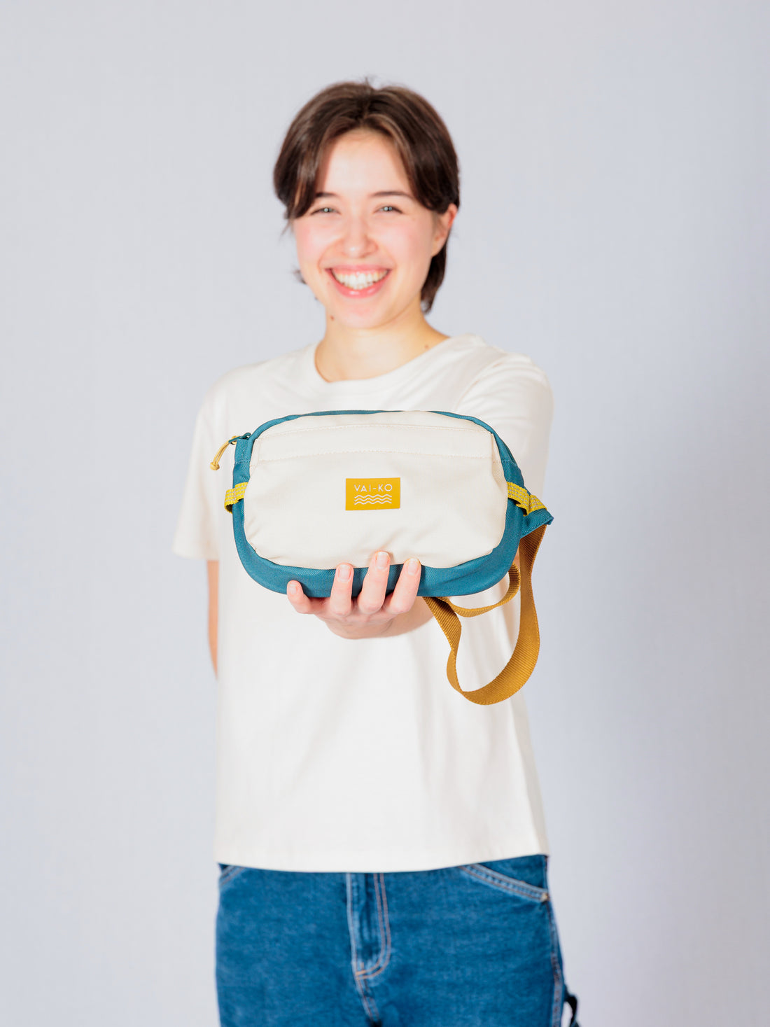 Person smiling and holding out a small beige and teal bag with a yellow label and strap.