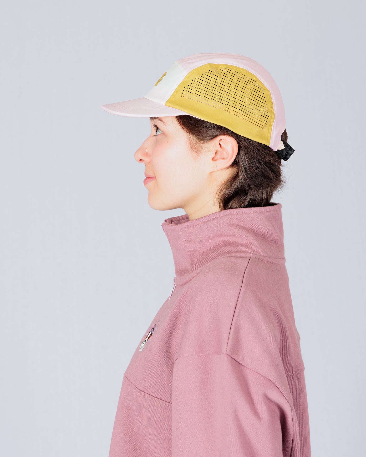 Person in a light pink and yellow cap and mauve jacket, shown in profile against a plain background.