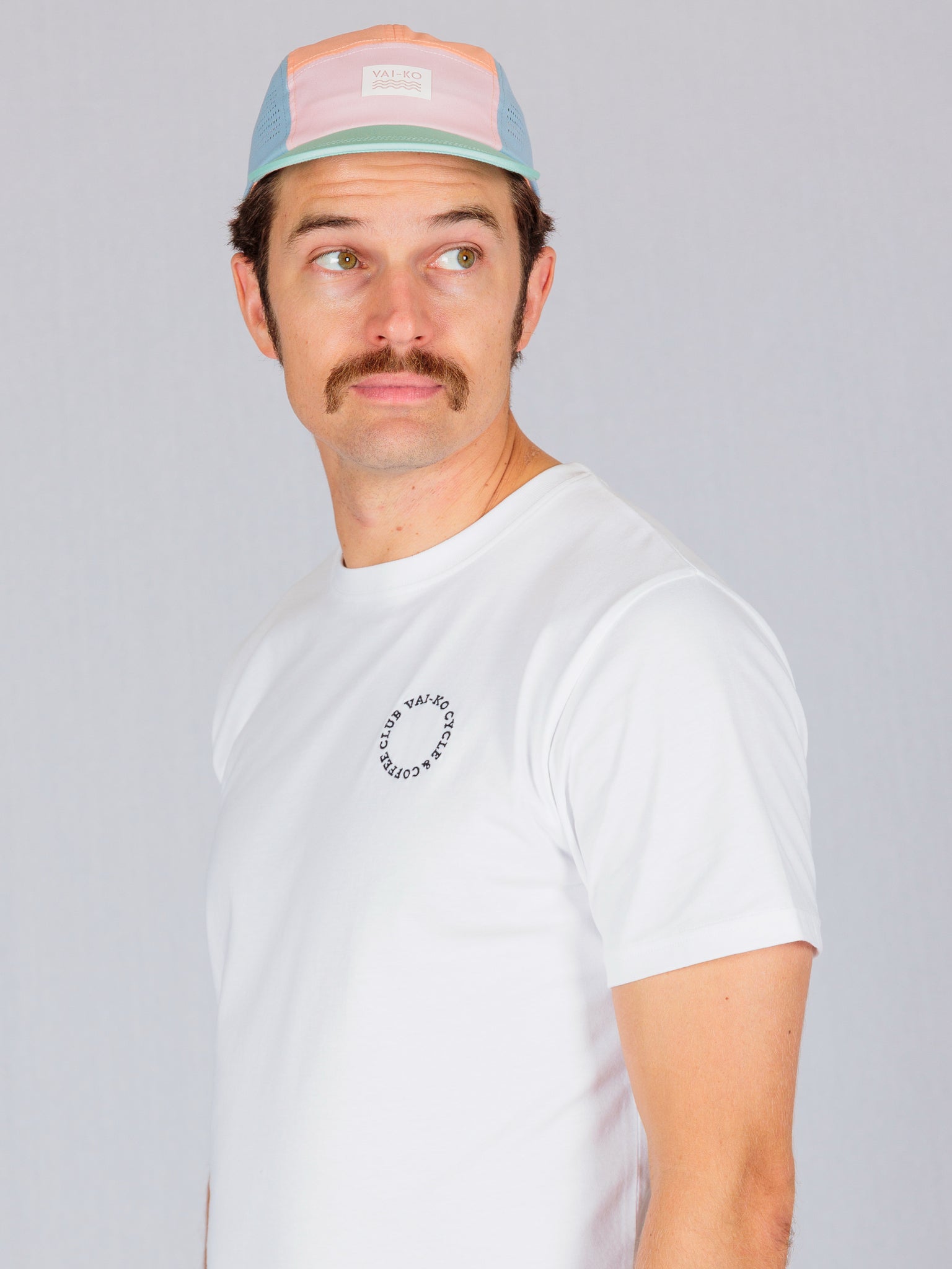 Man with a mustache wearing a pastel cap and white t-shirt, looking to his left against a plain background.