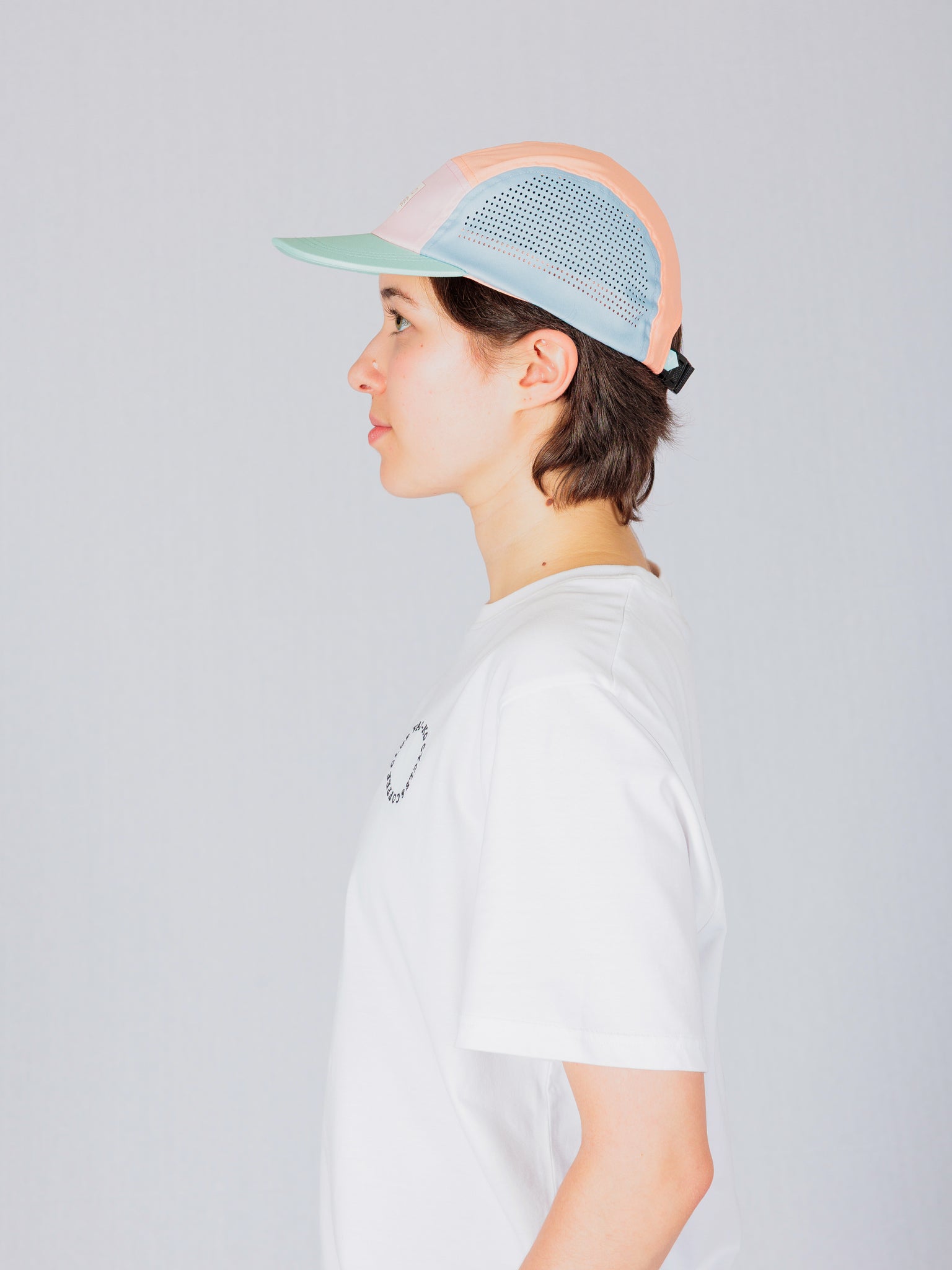Person in profile wearing a white shirt and a pastel-colored cap, standing against a plain light background.