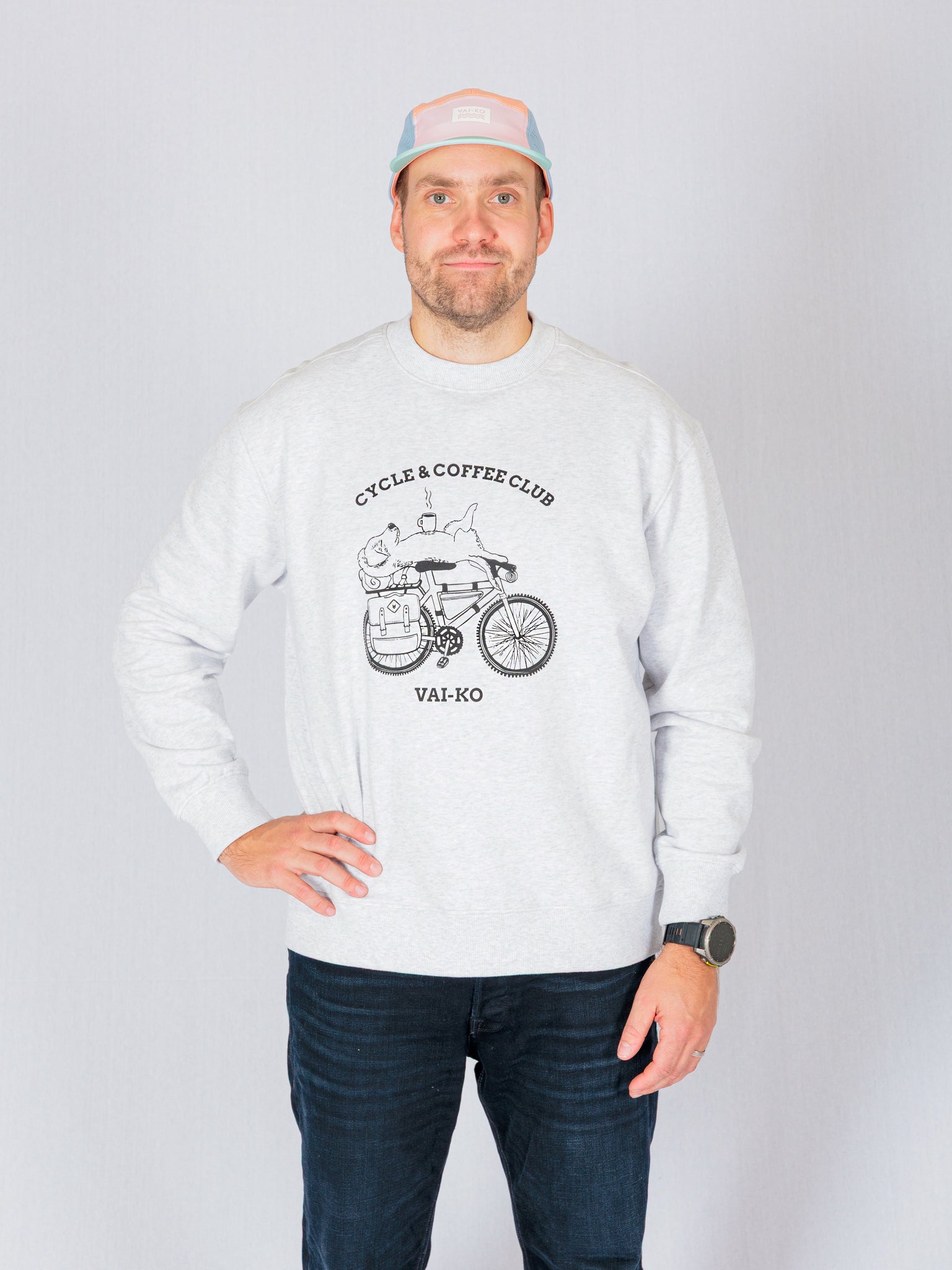 Man in a light sweatshirt with “Cycle & Coffee Club” graphic, wearing a pastel cap, stands against a plain background.