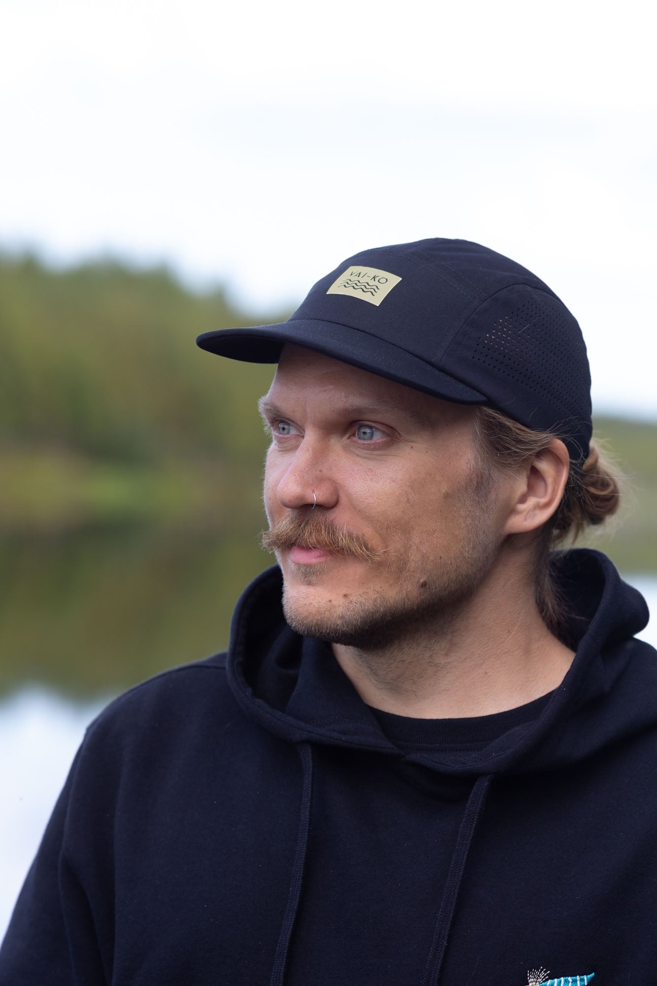 A man in a black hoodie and cap looks to the side outdoors with trees and water in the background.