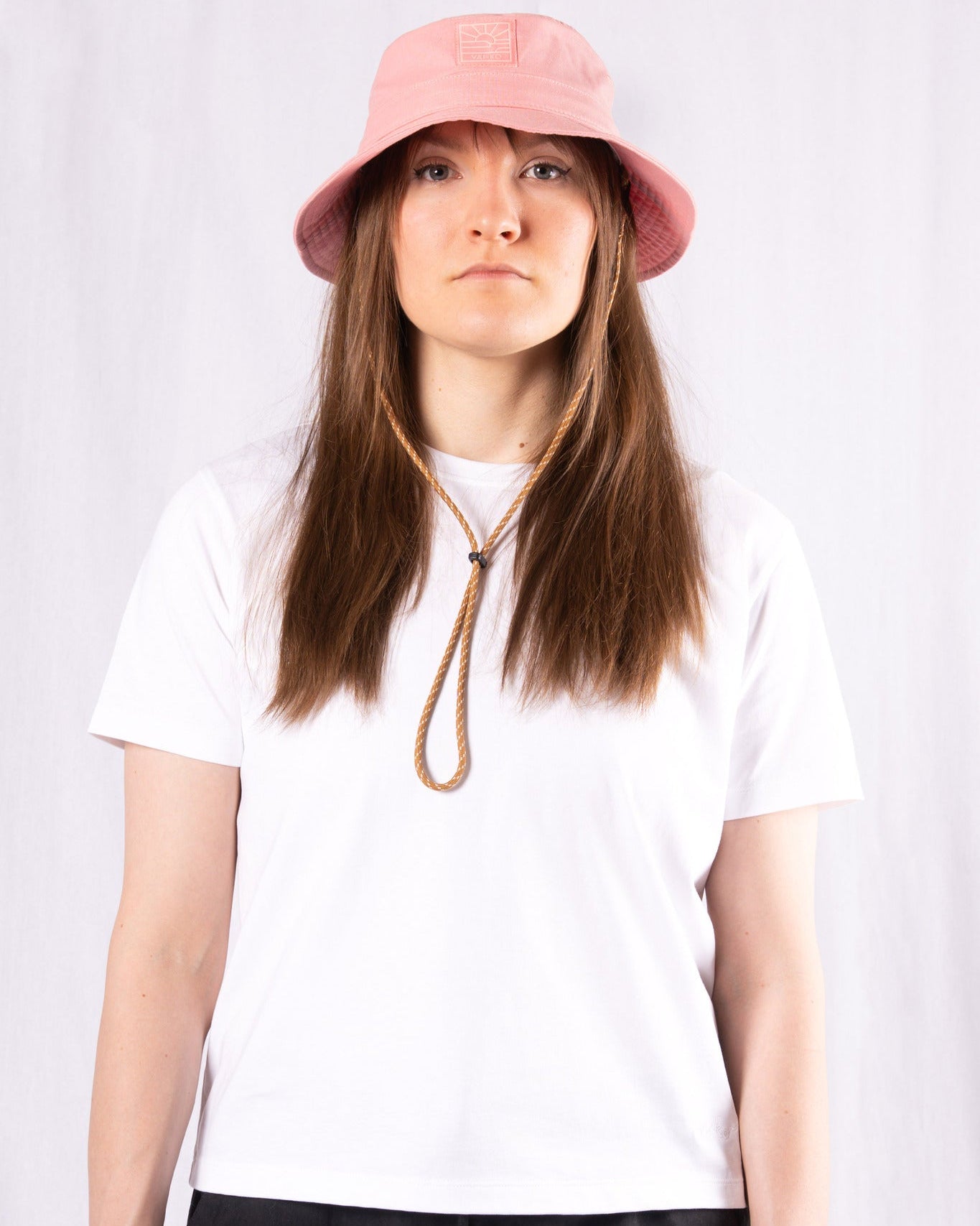 A person wearing a pink bucket hat, white t-shirt, and black pants stands against a plain white background.