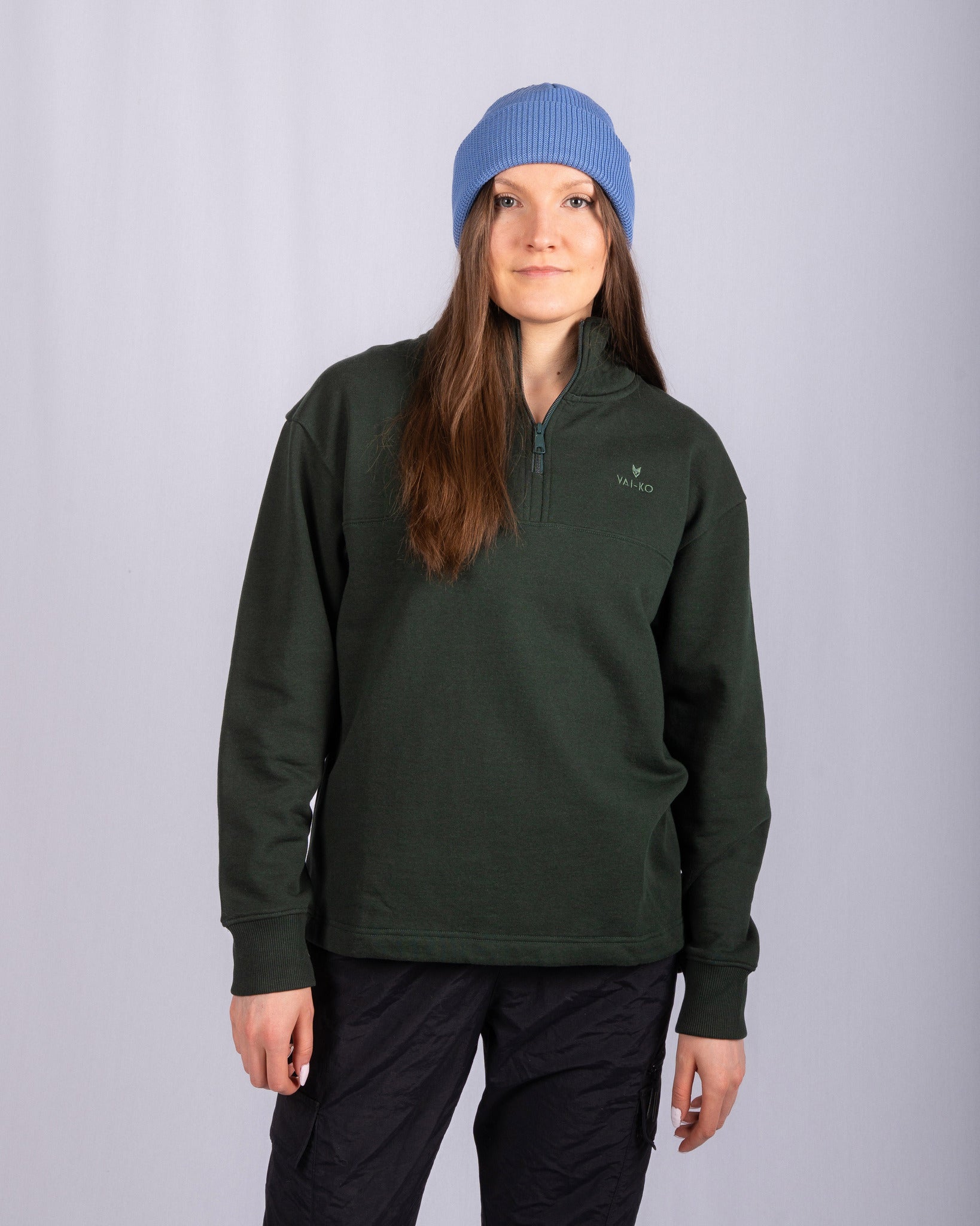 Woman in a blue beanie, dark green pullover, and black pants stands against a plain light background.
