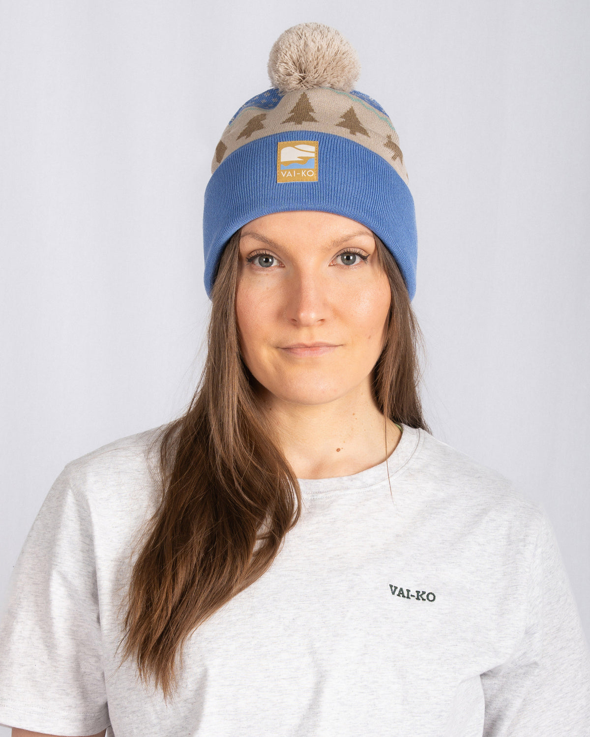 Woman in a blue and beige beanie with pom-pom, wearing a light gray VAI-KO t-shirt, facing forward.