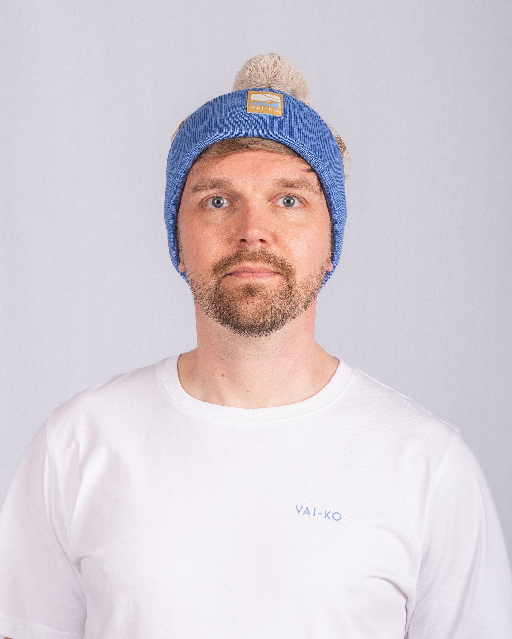 Man with a beard wearing a blue beanie and a white T-shirt stands against a plain light background.