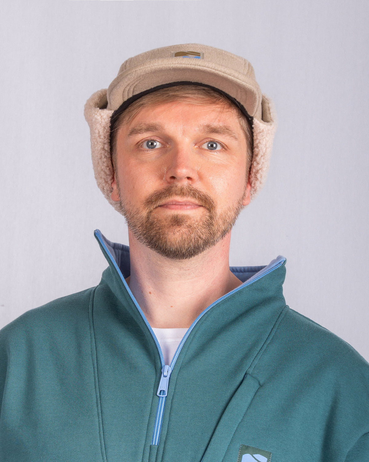 Man with beard wearing a tan sherpa ear-flap cap and teal zip-up jacket, facing camera against gray background.