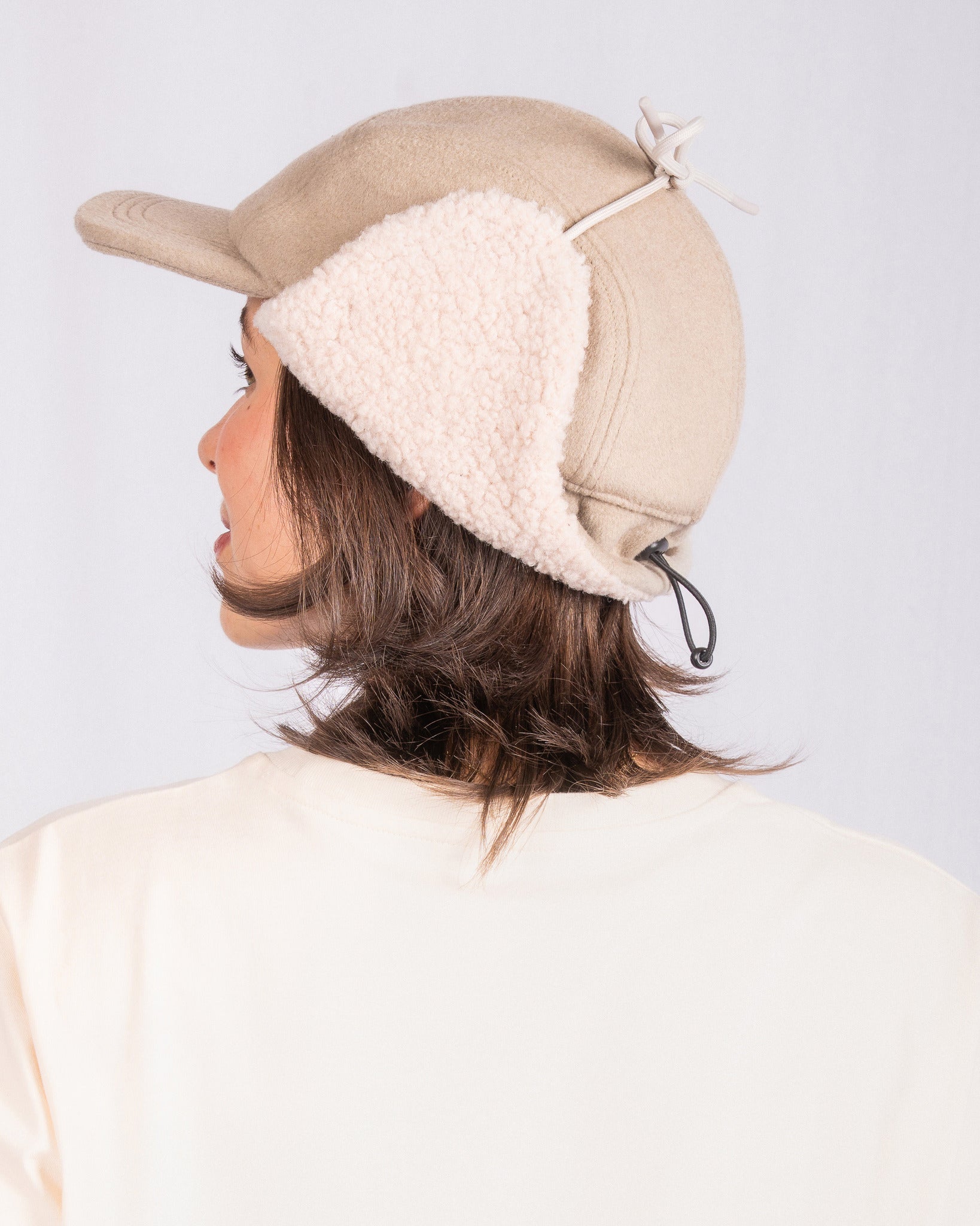 Person wearing a beige winter cap with fleece ear flaps, viewed from behind against a plain background.
