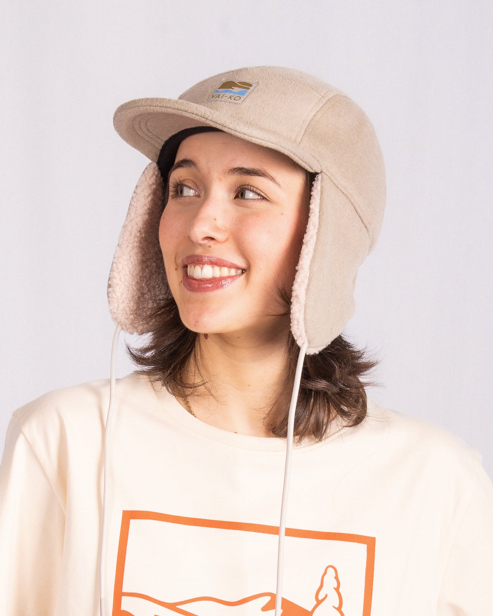 Smiling person in a beige winter hat with ear flaps and a cream shirt with an orange graphic.