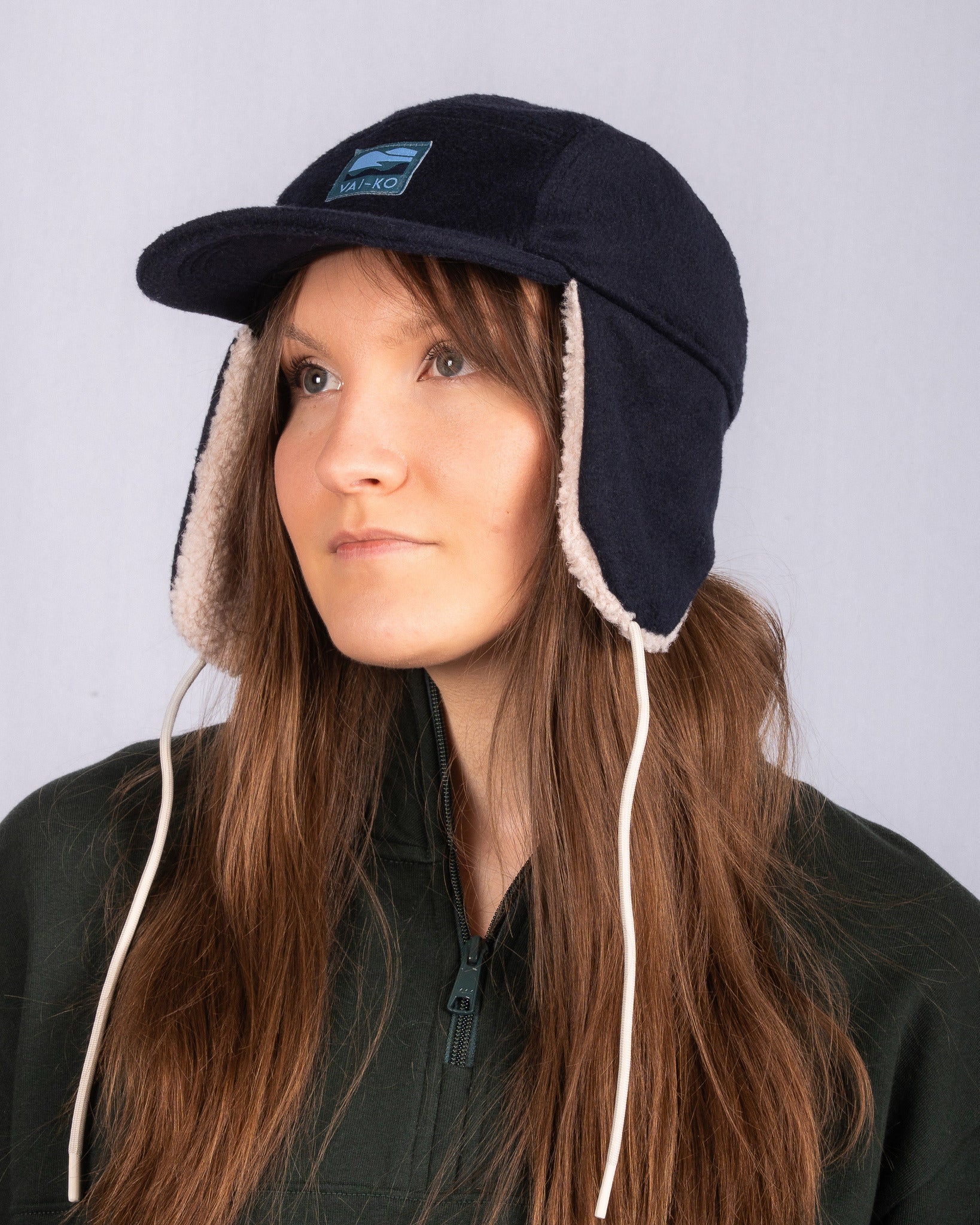 Woman in a dark trapper hat and green jacket looks upwards, against a plain light gray background.