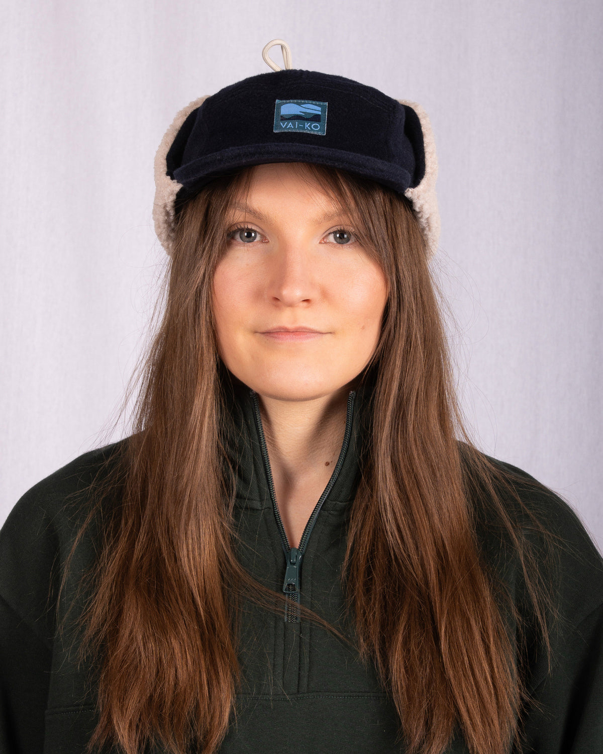 Woman with long brown hair wears a dark cap with ear flaps and a green zip-up jacket, facing forward.