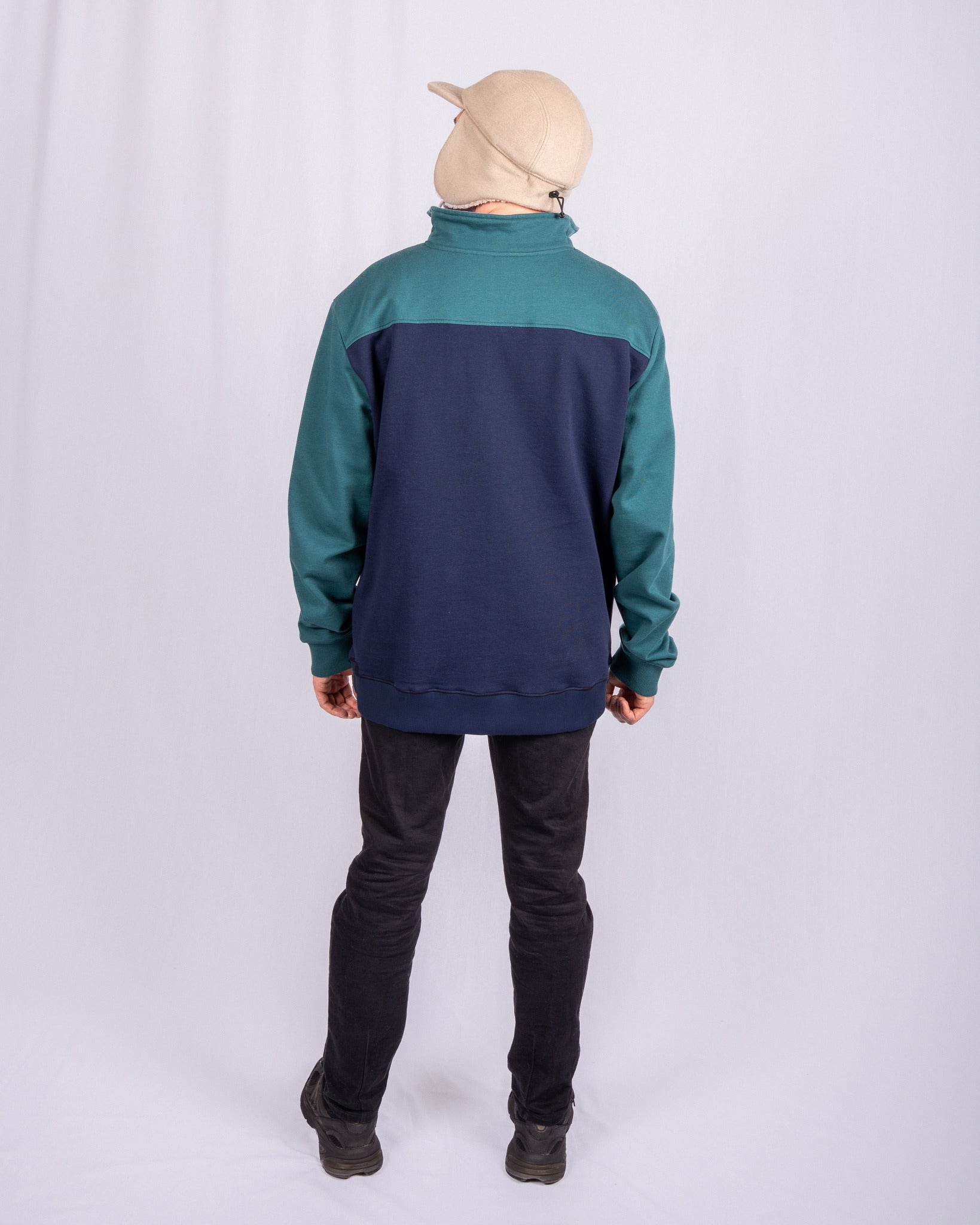 Person in a tan cap and blue-green sweatshirt stands facing away against a plain white backdrop.