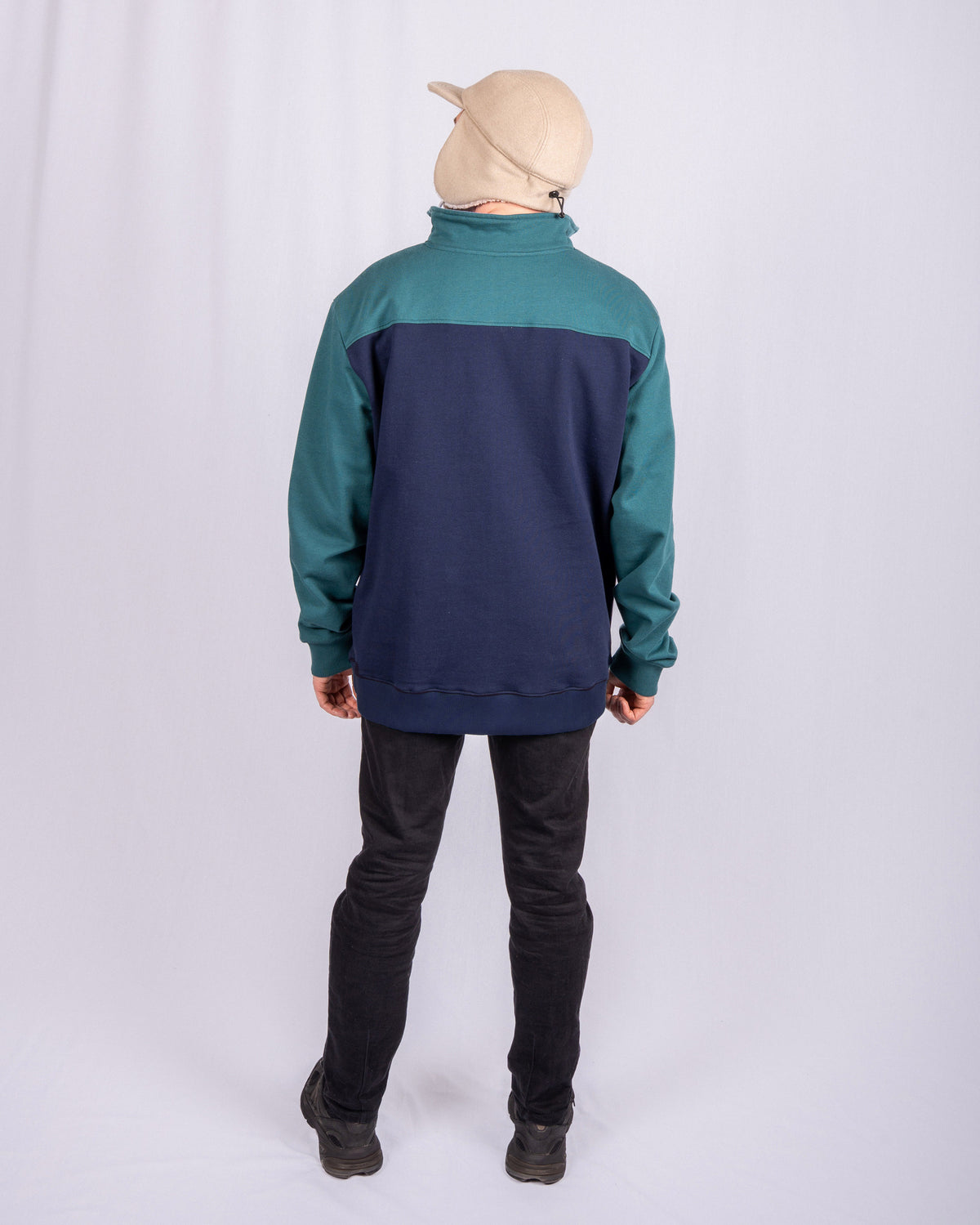 Person in a tan cap and blue-green sweatshirt stands facing away against a plain white backdrop.