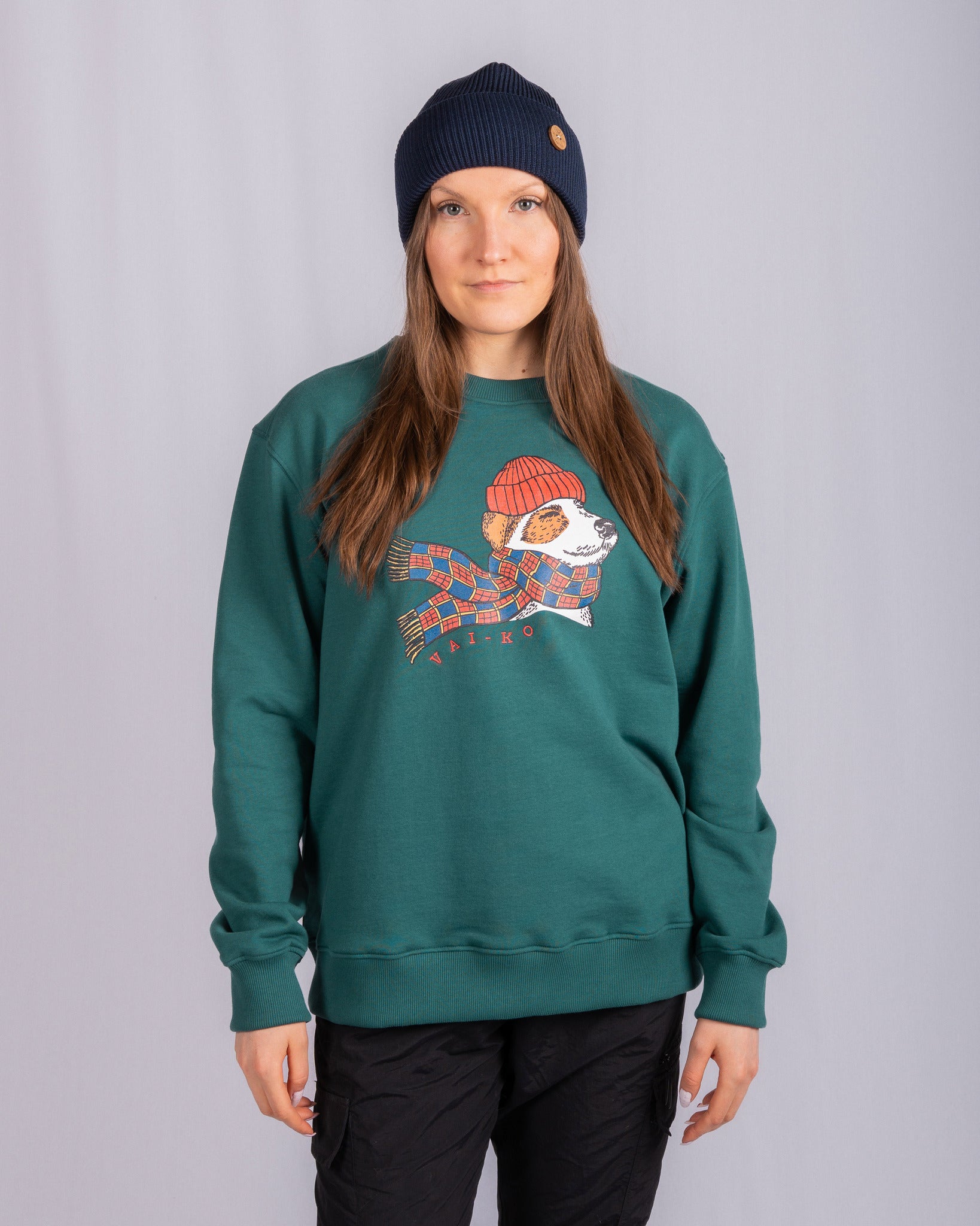 Woman in green sweatshirt with a cartoon graphic, black pants, and navy beanie, standing against gray background.