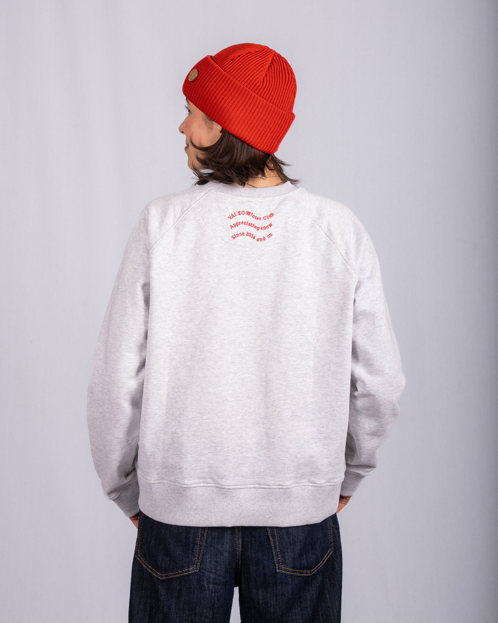 Person in red beanie and gray sweatshirt facing away, standing against a light gray background.