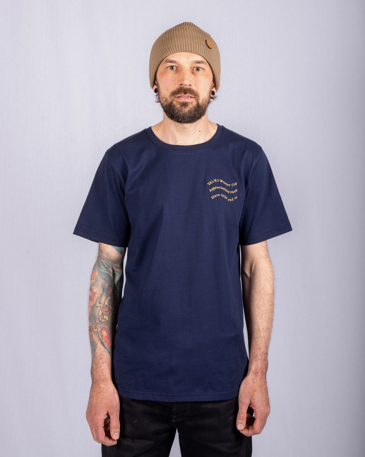 Man in navy t-shirt and tan beanie stands facing forward, showing tattooed arm, against plain background.