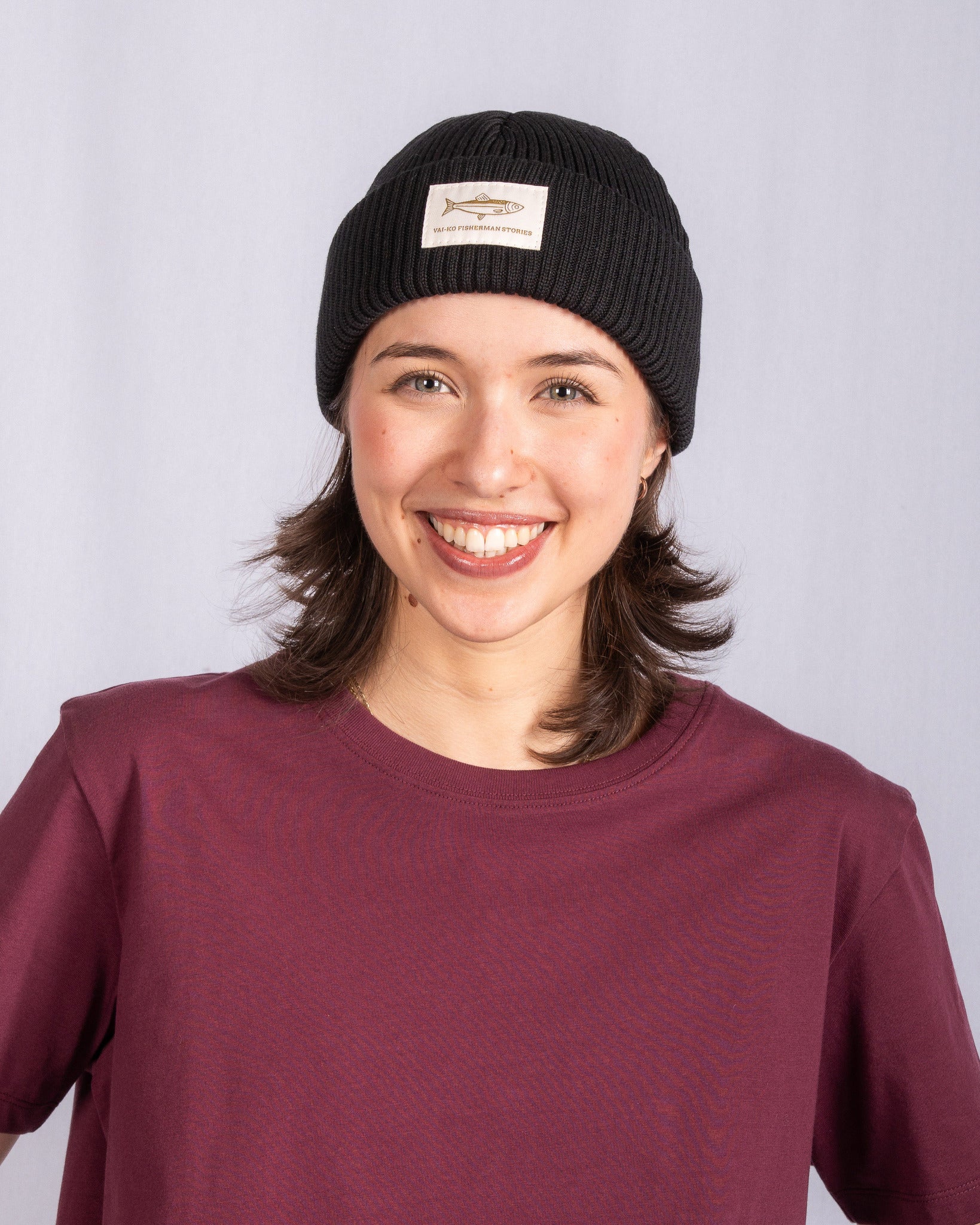 Smiling person in a maroon shirt and black beanie with a fish patch, standing against a light background.