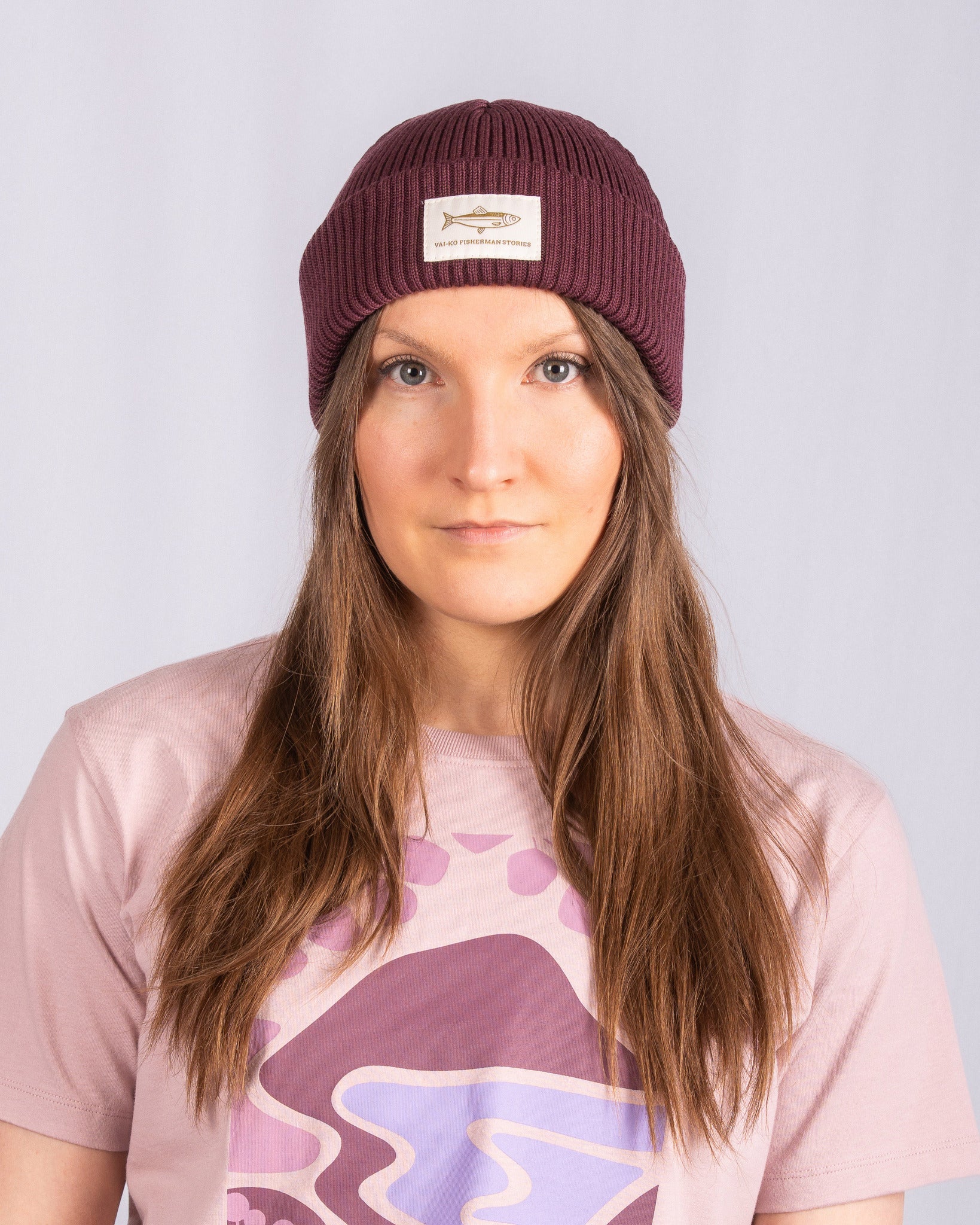 Woman with long brown hair wearing a maroon beanie and a pink graphic t-shirt, facing forward.