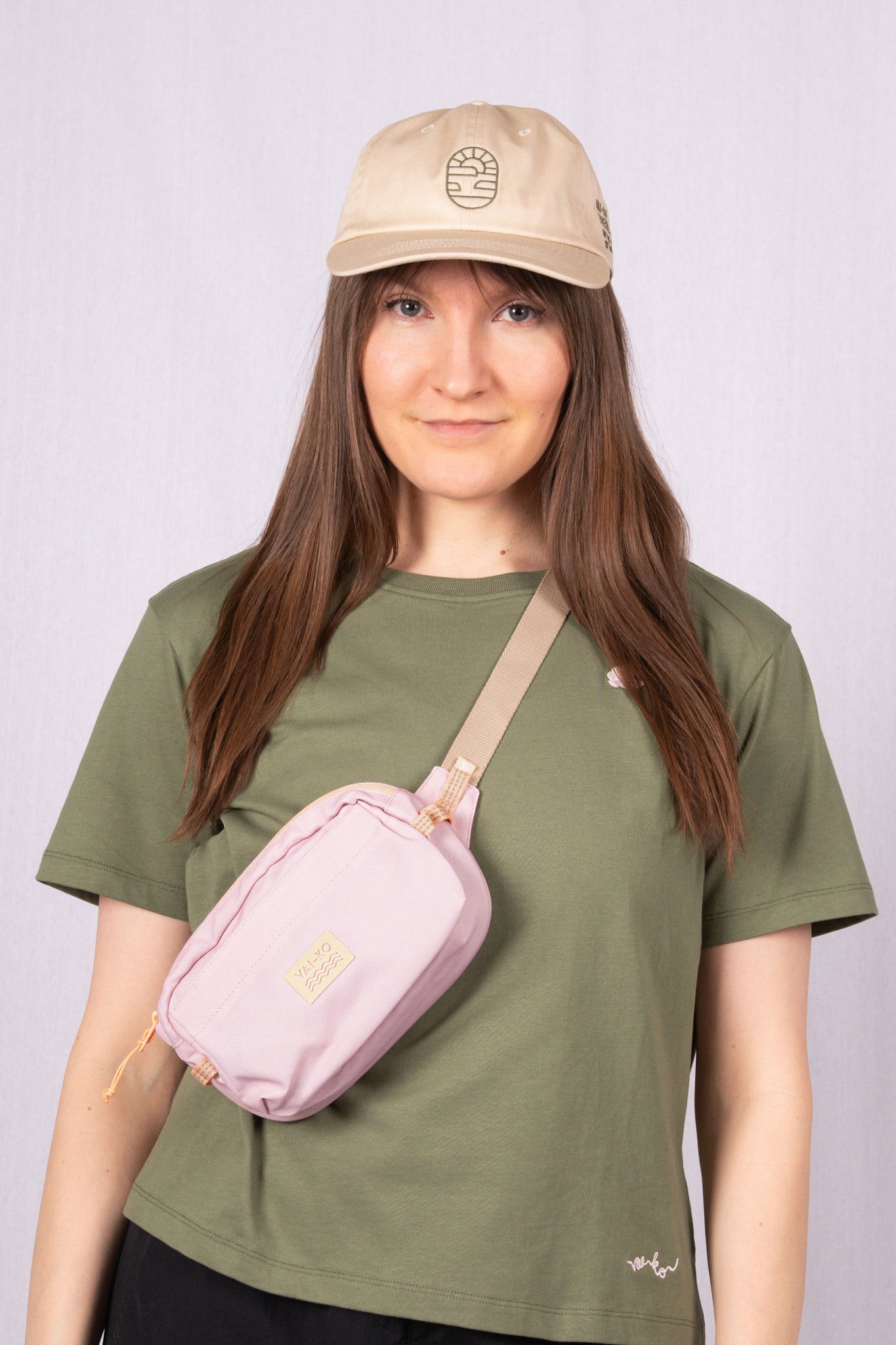 Woman in a khaki cap, olive green t-shirt, and pink crossbody bag, standing against a light background.