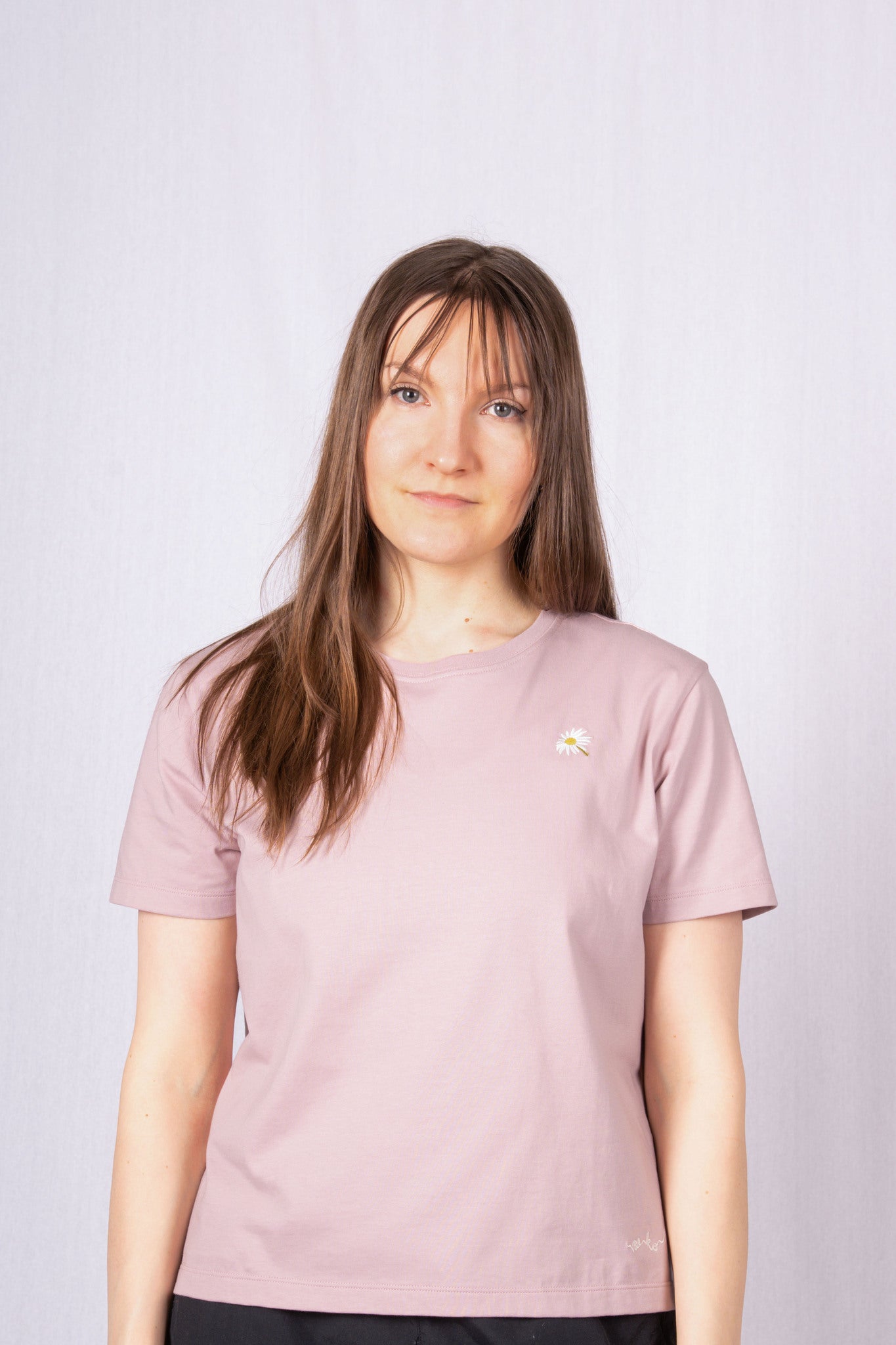 Woman with long brown hair wearing a mauve t-shirt, standing against a plain light background.