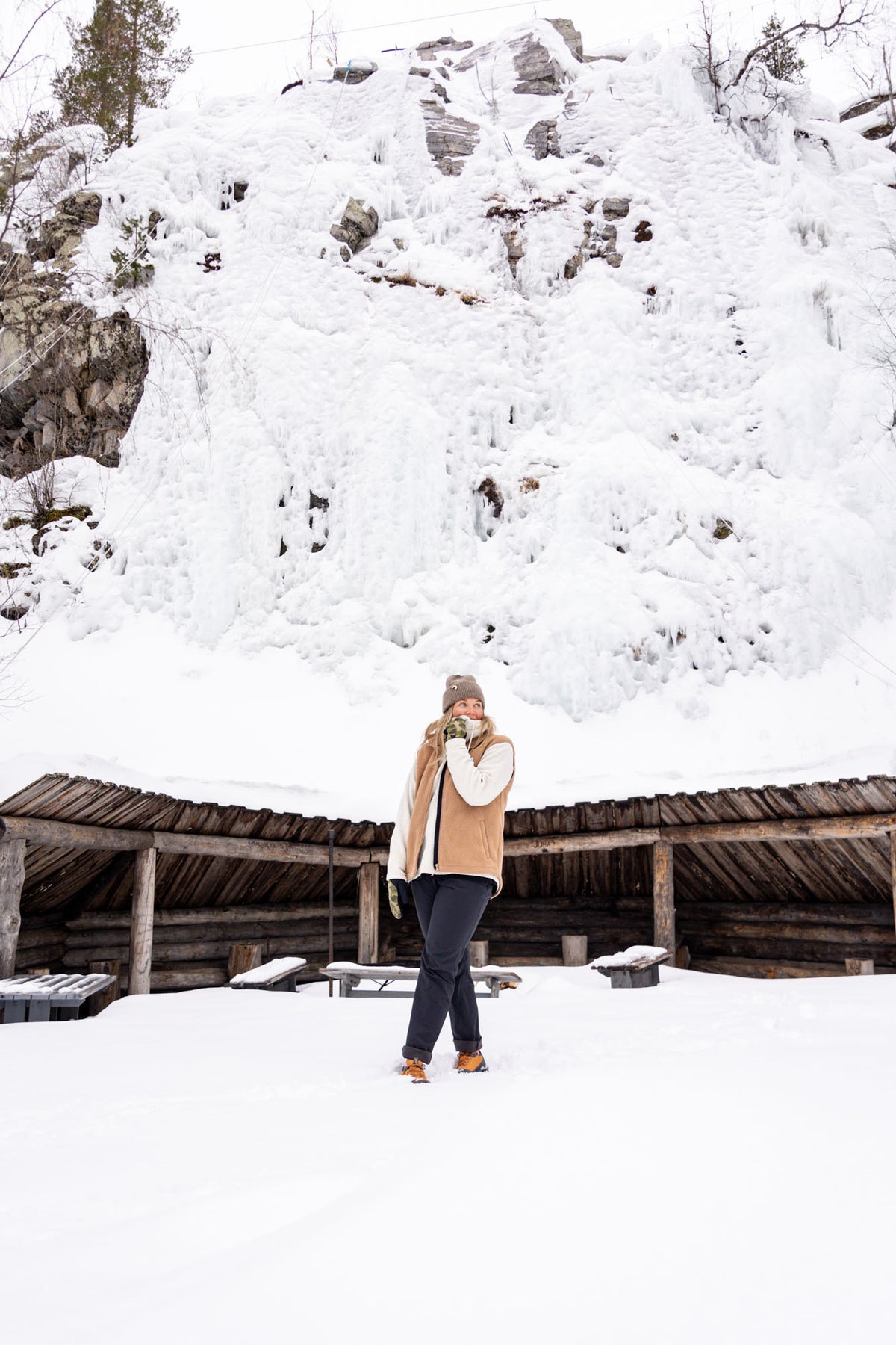 Person standing in snow, wearing winter clothes, with a snowy rock cliff and wooden shelter in the background.