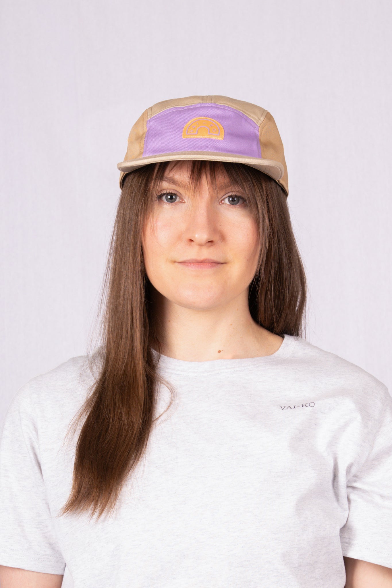 Woman with long brown hair wearing a beige and purple cap and a light gray t-shirt, facing forward.
